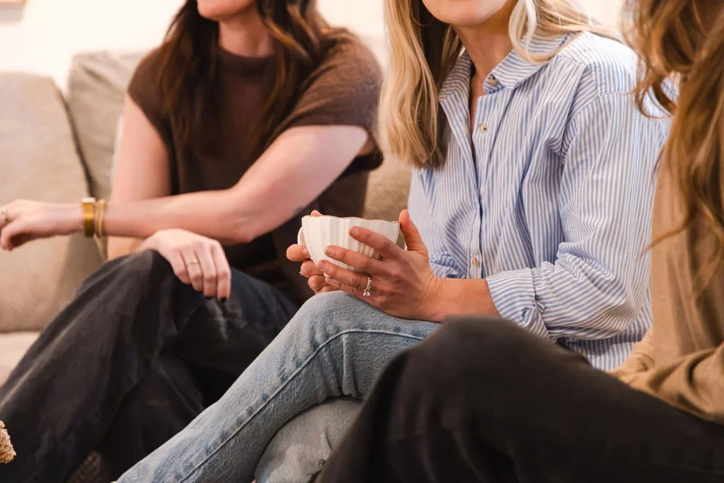 Three women sitting on a beige couch having a conversation, one woman holds a white ceramic mug.