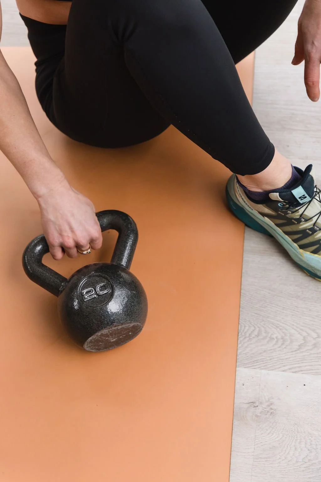 Person sitting on an exercise mat holding a 20-pound kettlebell with one hand, wearing black leggings and athletic shoes.