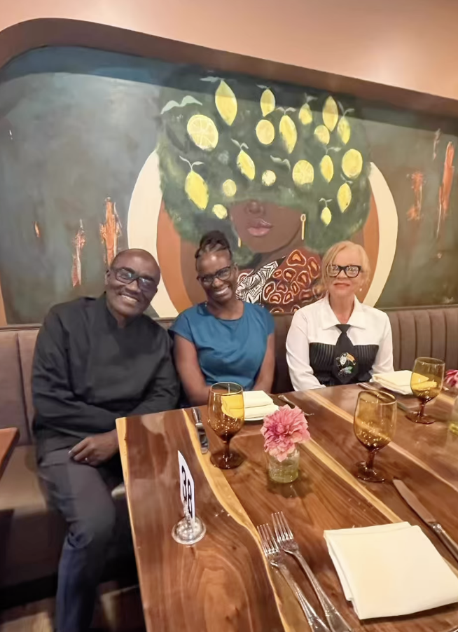 Three people sitting at a restaurant table with drinks and napkins, smiling; large artwork of a woman with large yellow lemon-shaped hair hanging on the wall behind them.