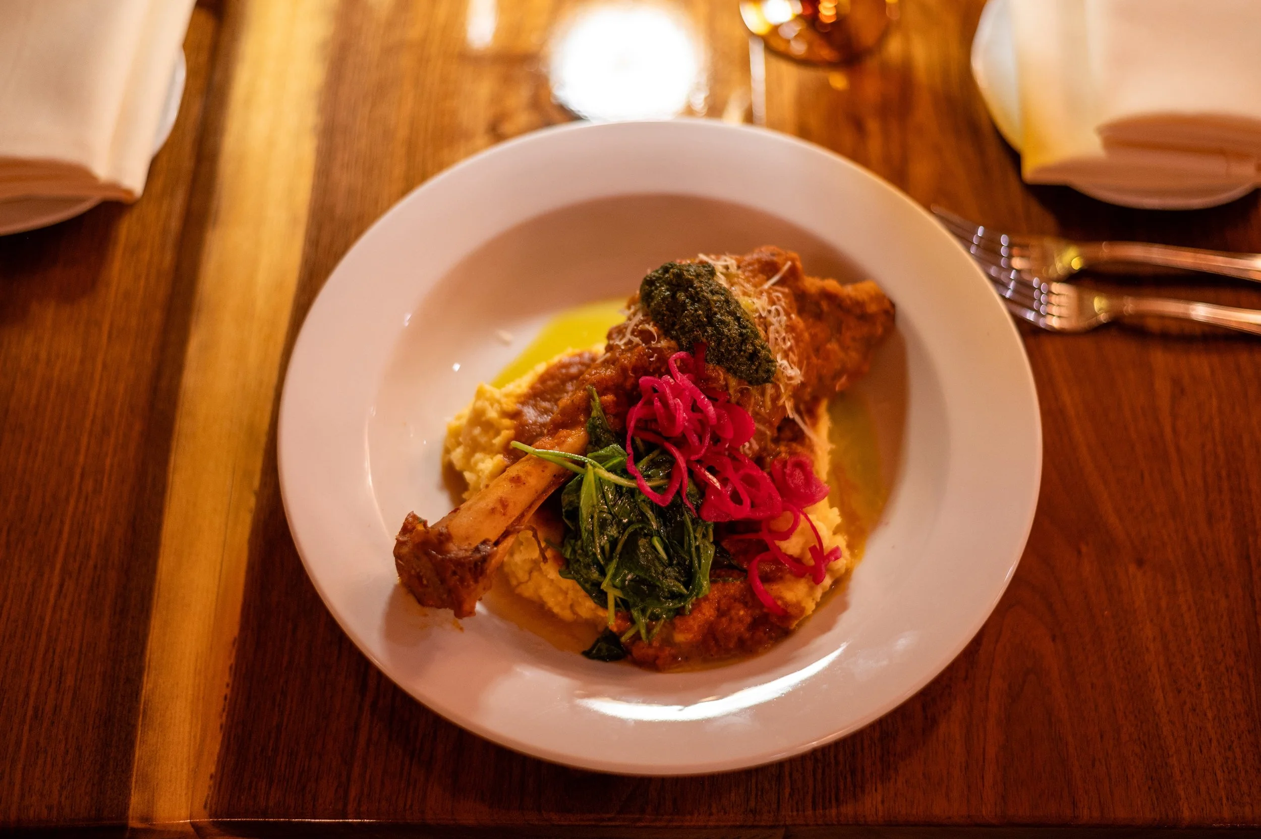A white plate with a serving of mashed potatoes topped with gravy, green leafy vegetables, a piece of toasted bread, and garnished with pickled red onions and grated cheese, placed on a wooden table with napkins and cutlery.
