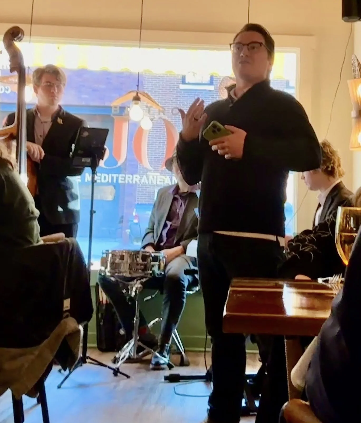 A man with glasses and dark hair holding a phone, standing in a restaurant or cafe, speaking or singing, with several people seated and listening, and a musician with a drum set in the background.