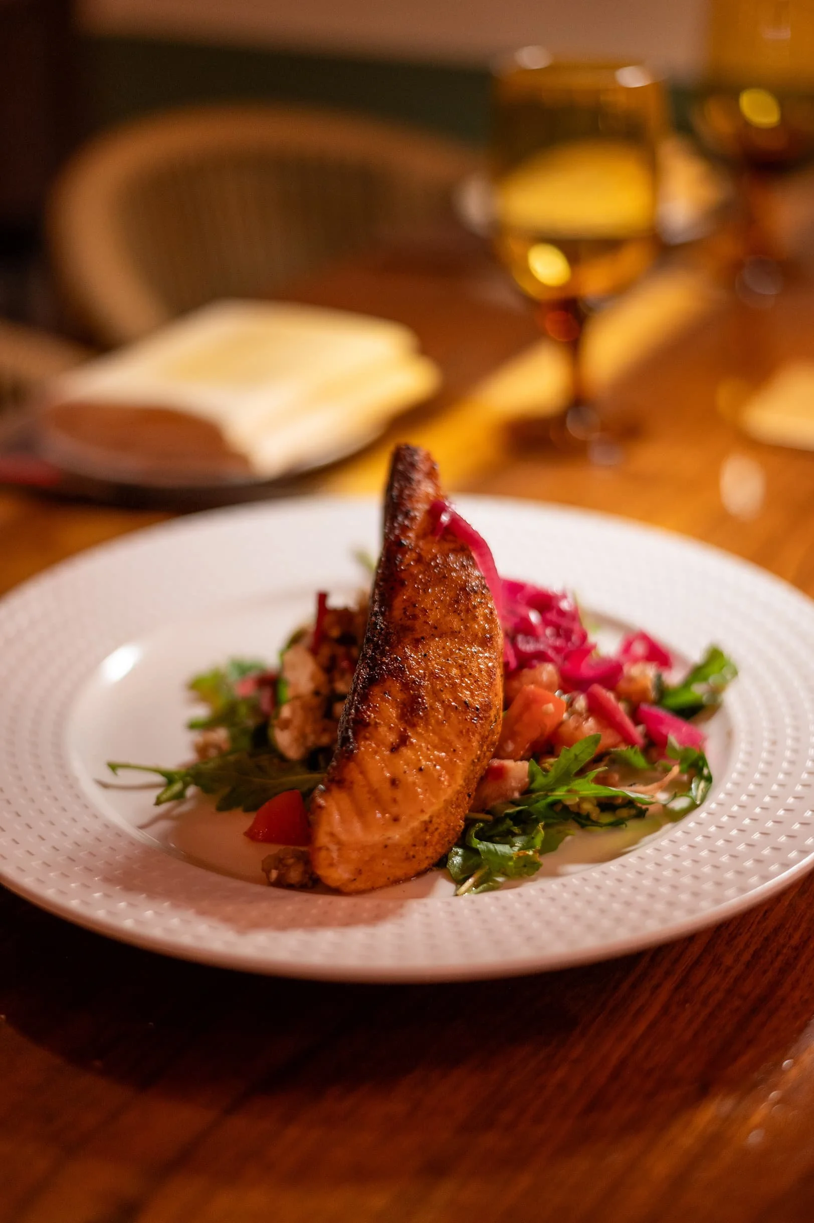 Plated dish featuring a piece of cooked salmon on a bed of mixed greens, chopped vegetables, and pink pickled onions, served on a white plate with a textured rim, with a blurry background of wine glasses and utensils.