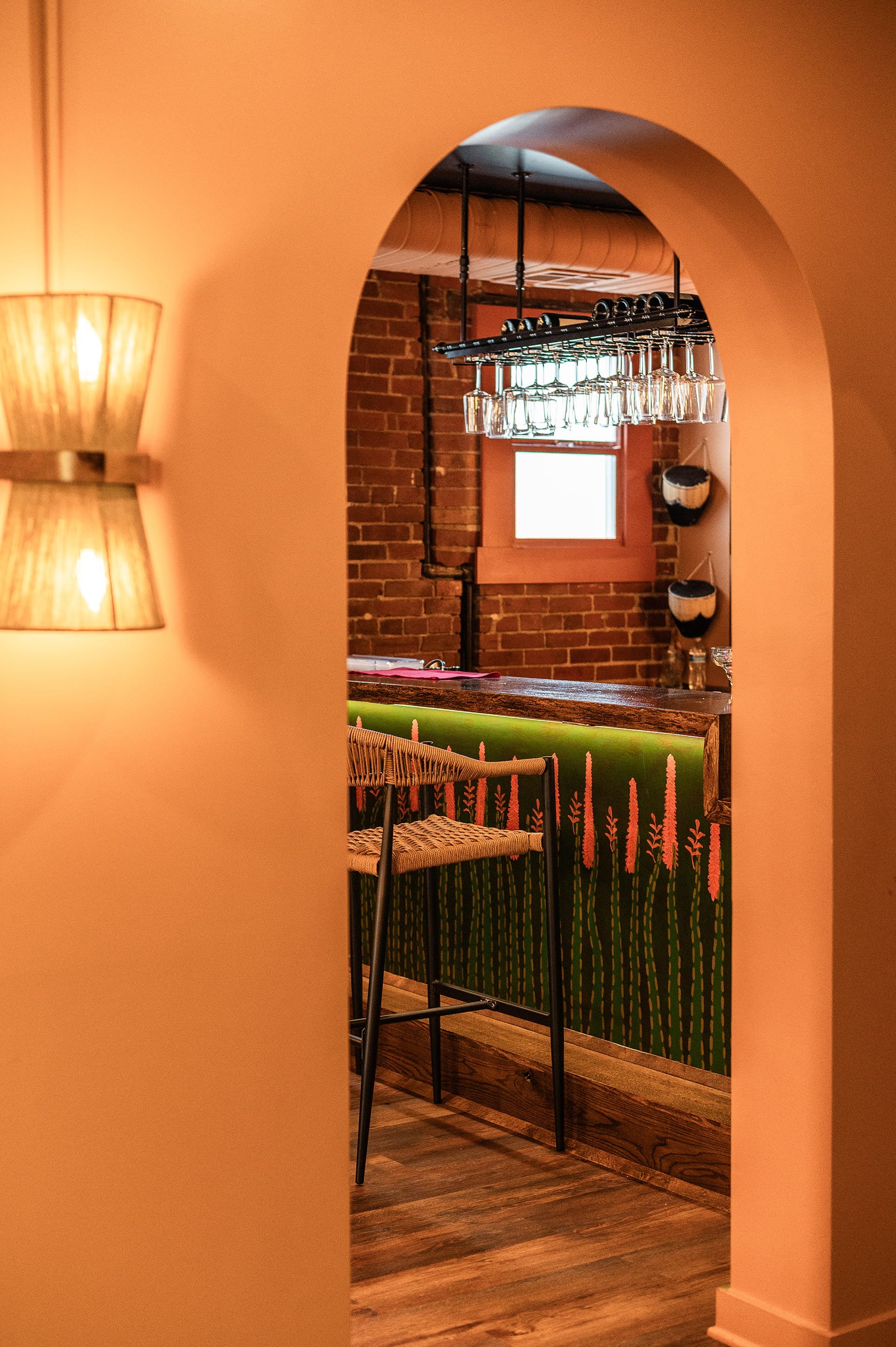 View through a wall opening showing a bar area with a hanging glass rack, brick wall, small window, and decorative pottery against the wall.
