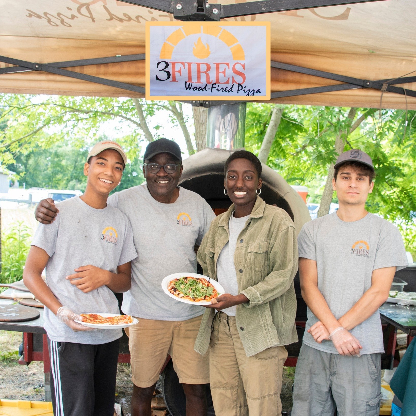 Four people standing outdoors under a tent, one of whom is holding a pizza; a sign above them reads '3 Fires Wood-Fired Pizza'.