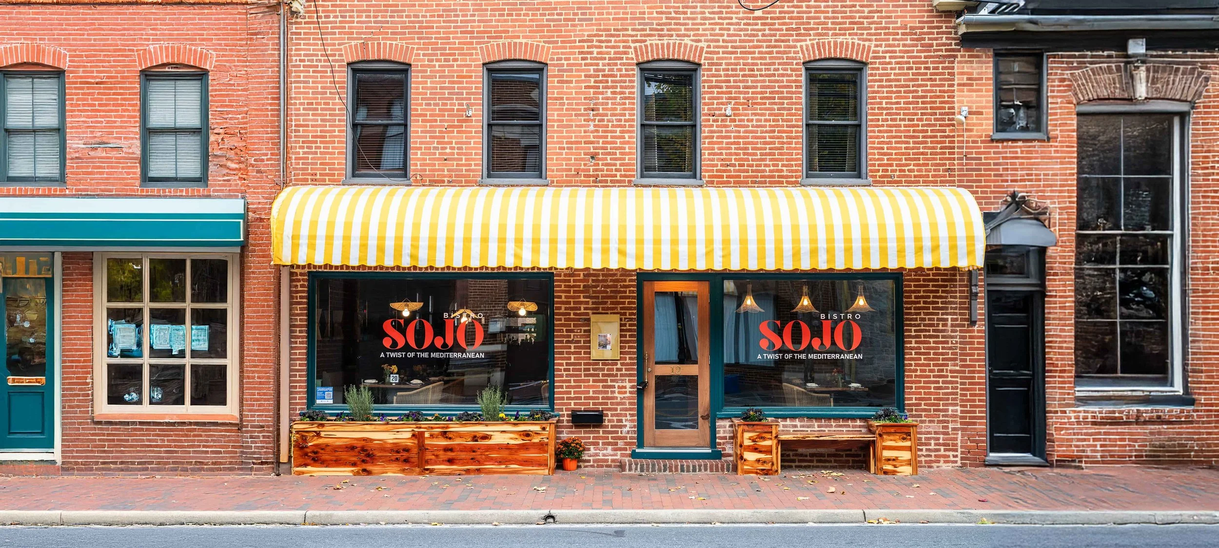 Street view of a SOJO Bistro. The restaurant has large windows, a wooden door, and a yellow and white striped awning. There are some potted plants and small flower boxes outside.