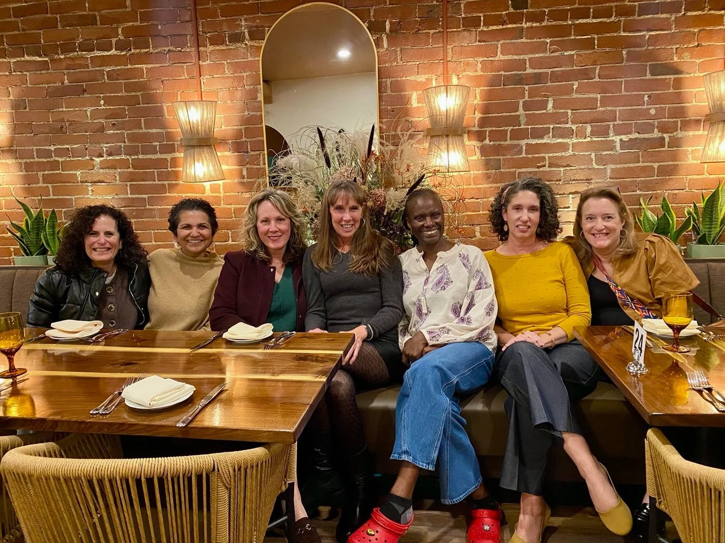 Seven women sitting at a restaurant table in front of a brick wall and a mirror, smiling for the camera, with table settings and plants in the background.