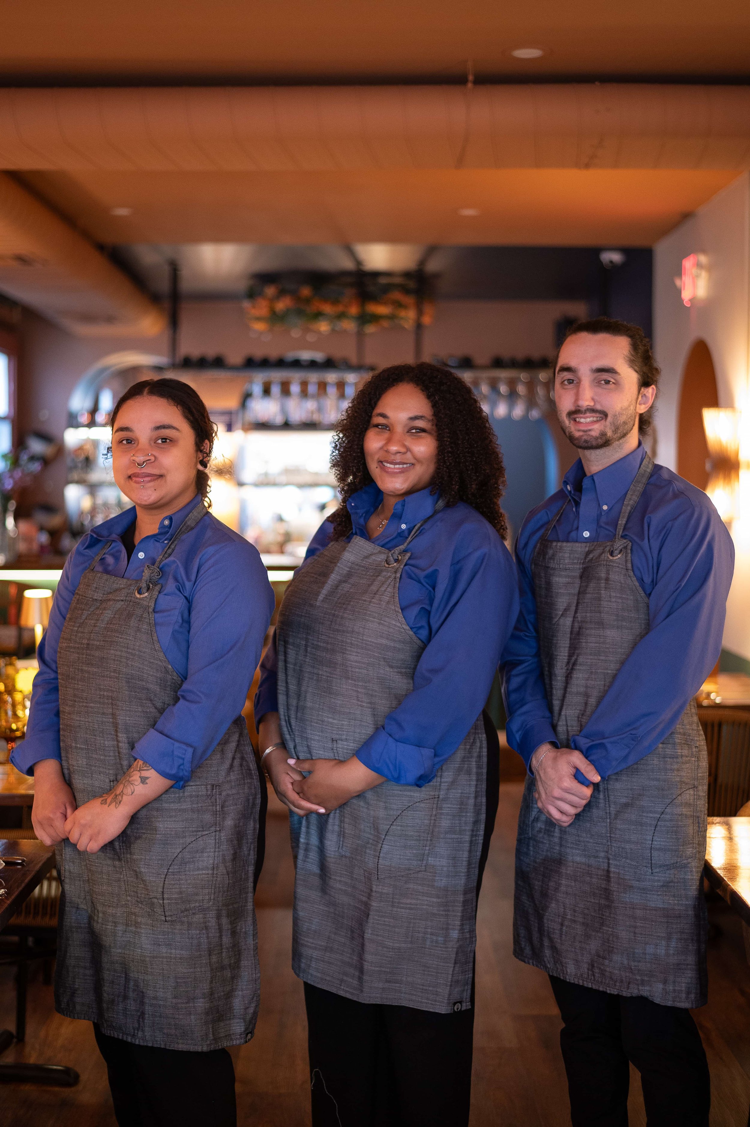 Three restaurant staff members standing together in a warmly lit dining area, wearing blue shirts and gray aprons, smiling at the camera.