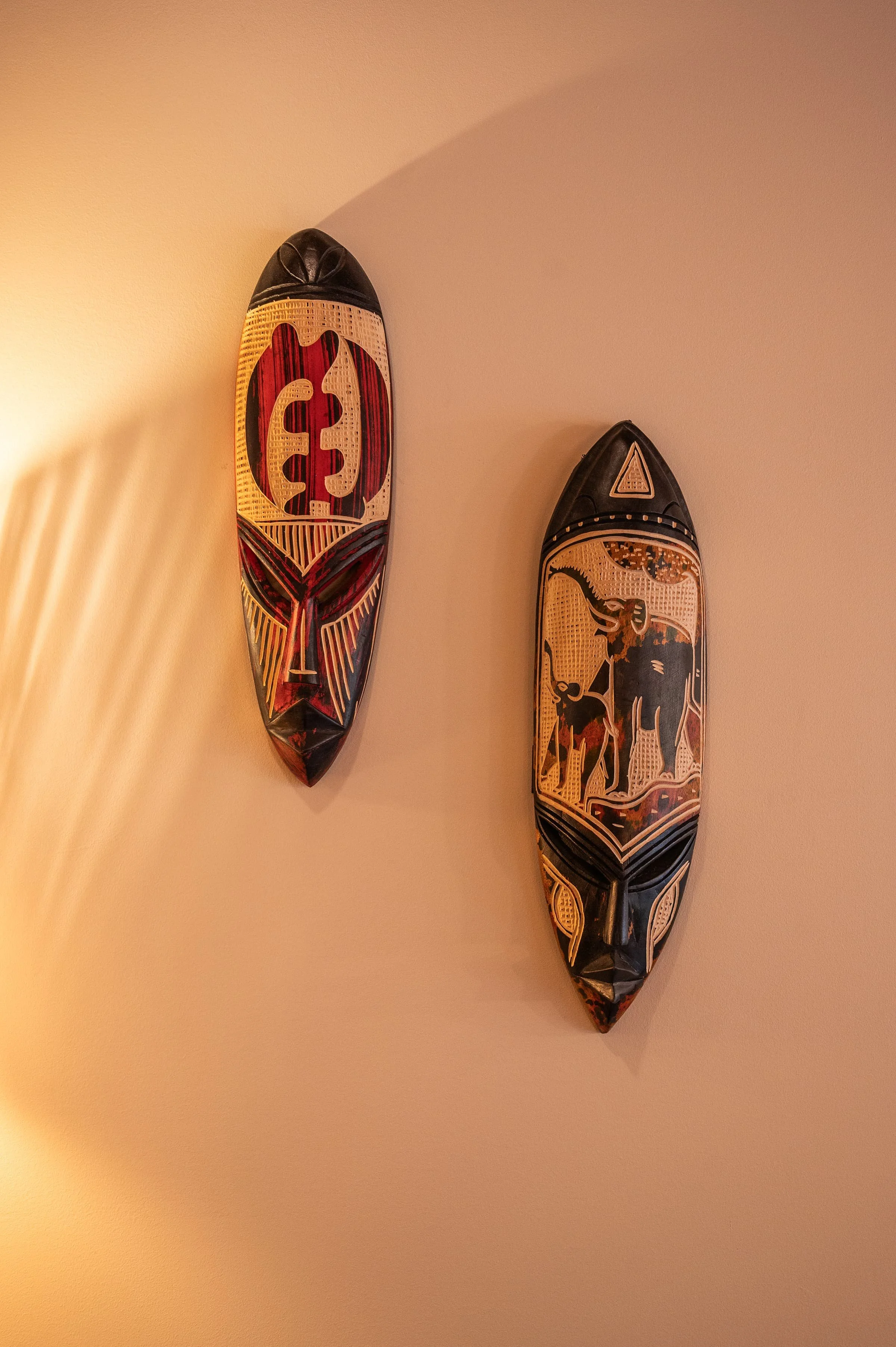 Two decorative tribal masks mounted on a beige wall, one with red and black patterns and the other with a dolphin and other design elements.
