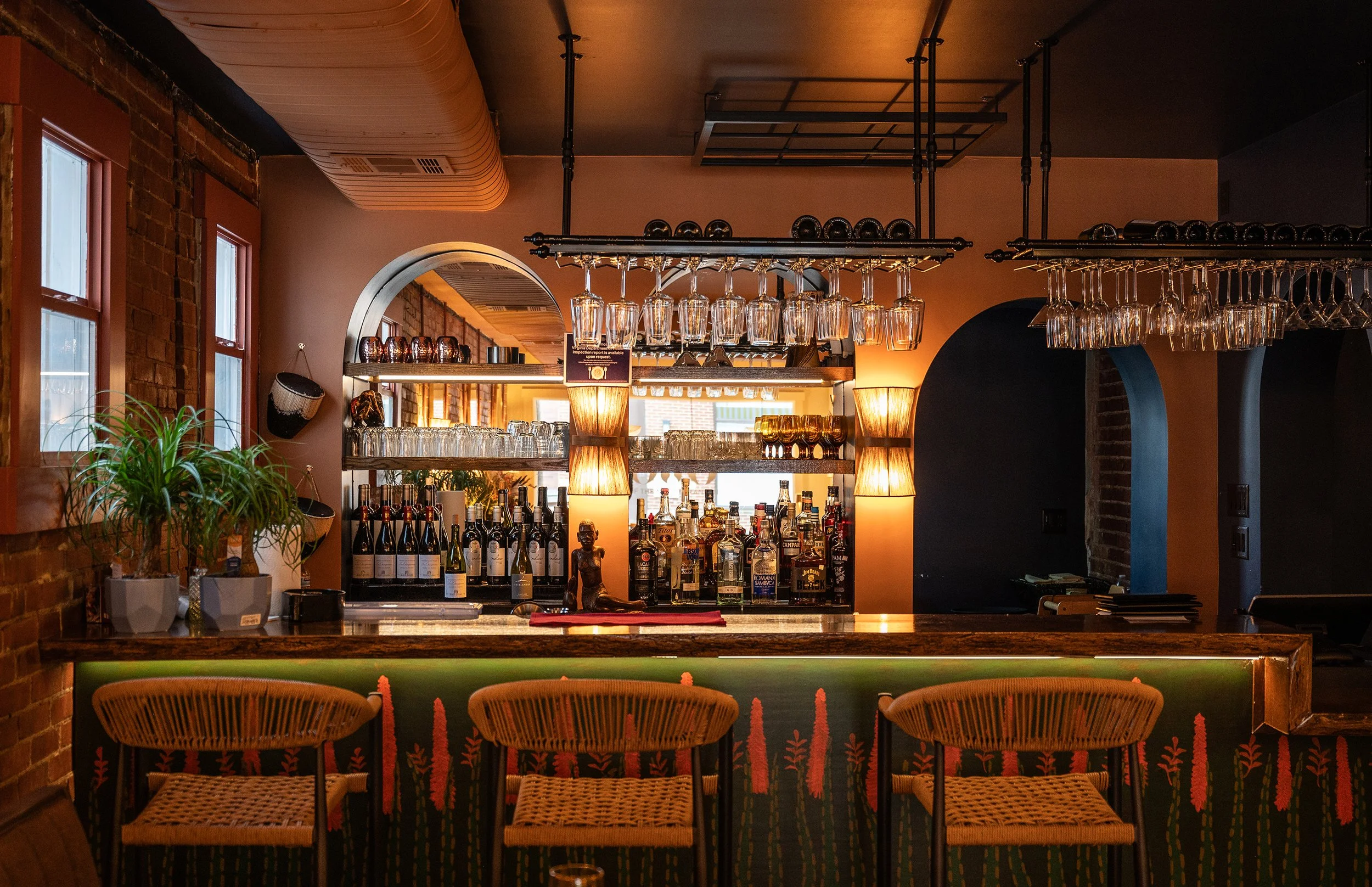 The bar area with a dark ceiling, brick walls, and windows. Hanging glasses above the counter and bottles of alcohol arranged on shelves. Three wicker bar stools with backs are at the counter, which has a decorative curtain with a floral pattern.