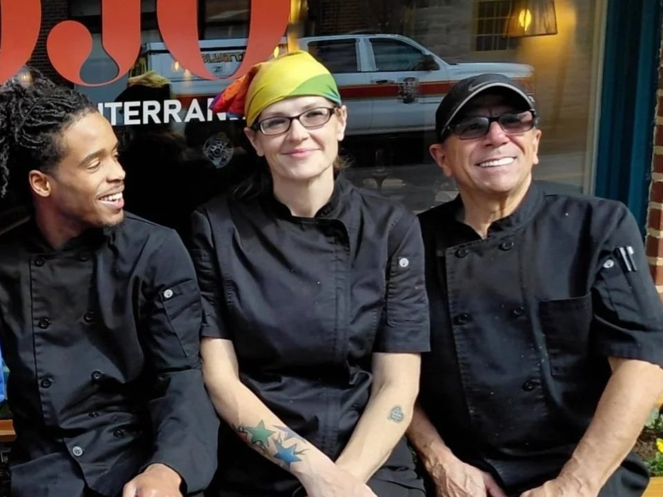Three chefs in black uniforms sitting outside a restaurant, smiling and talking to each other. The woman in the middle wears glasses and a colorful headscarf.