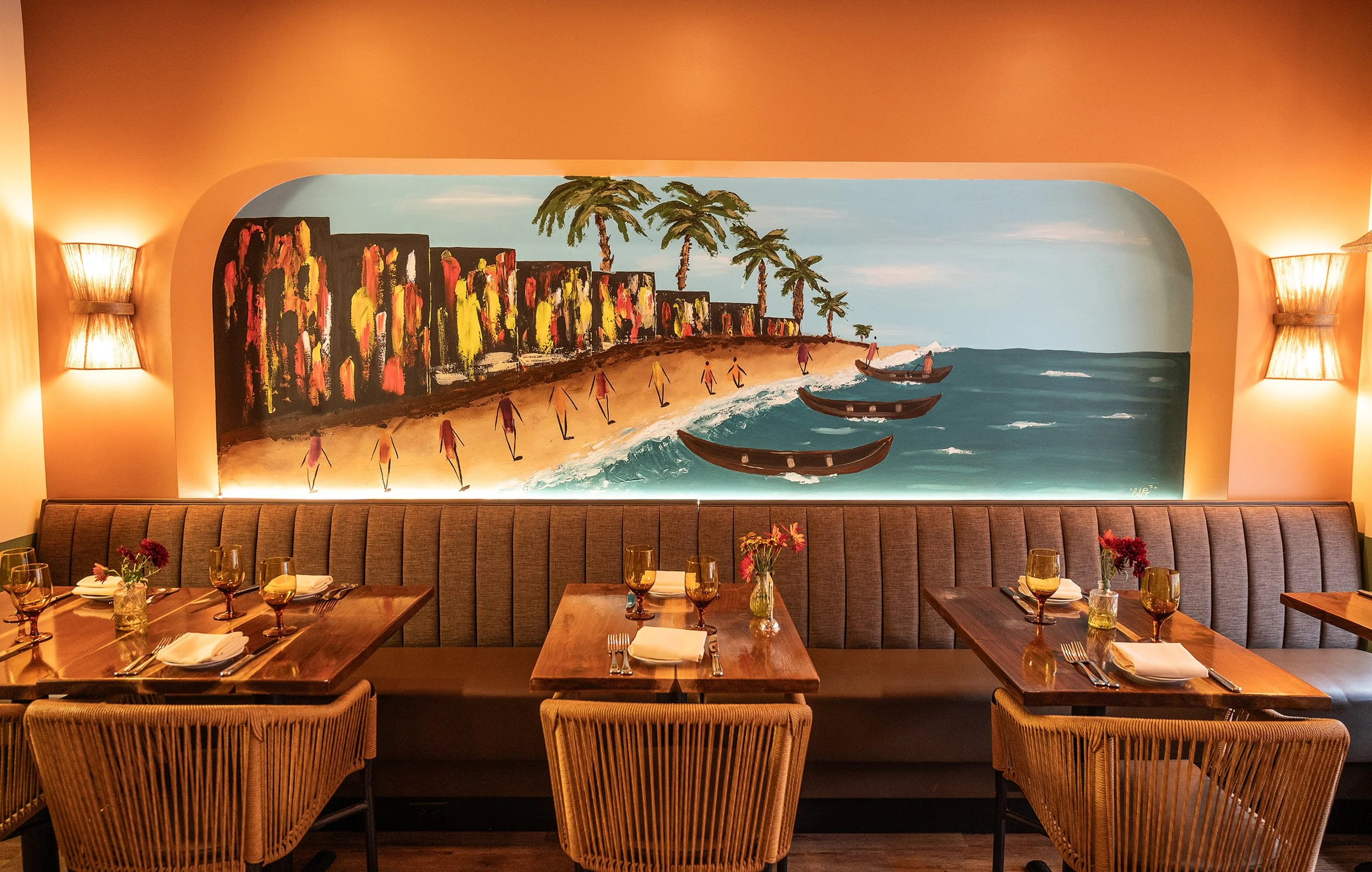 A restaurant interior with wooden tables set with wine glasses, white napkins, and small flower arrangements. A large, colorful painting of a beach scene with palm trees, boats, cliffs, and people decorates the wall behind the seating area, flanked by two wall sconces emitting warm light.