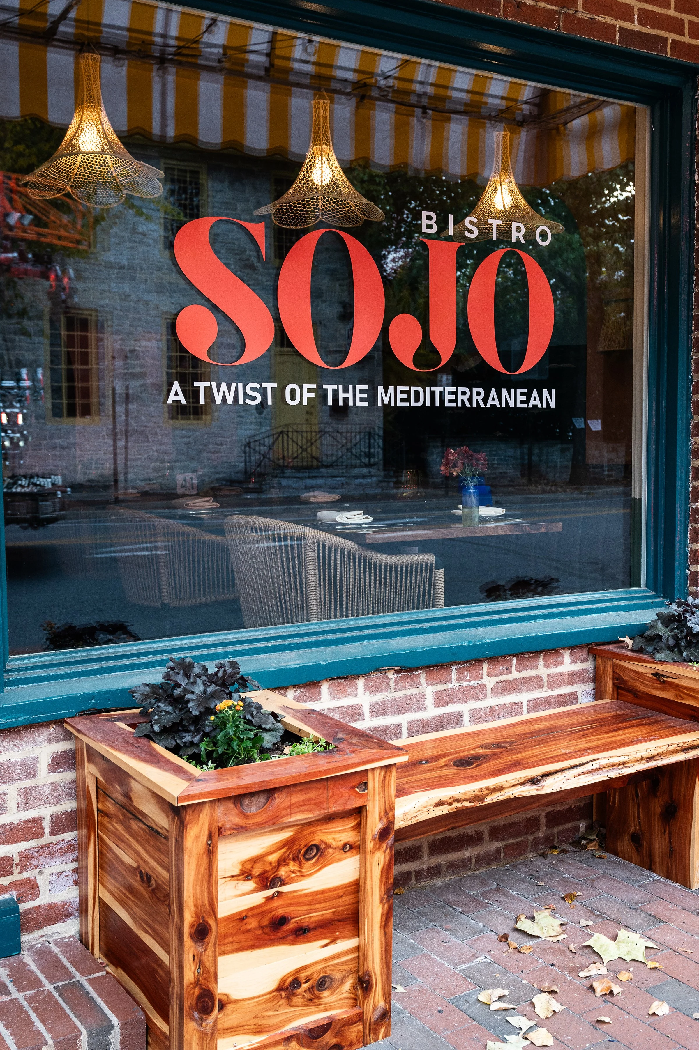 A window display advertising a bistro offering a $0.00 menu, with the tagline 'A Twist of the Mediterranean.' Inside, there are hanging lamps, tables set with plates and napkins, and a single blue vase with pink flowers. Outside, a wooden planter with purple and green plants is on a brick sidewalk, with fallen leaves scattered about.