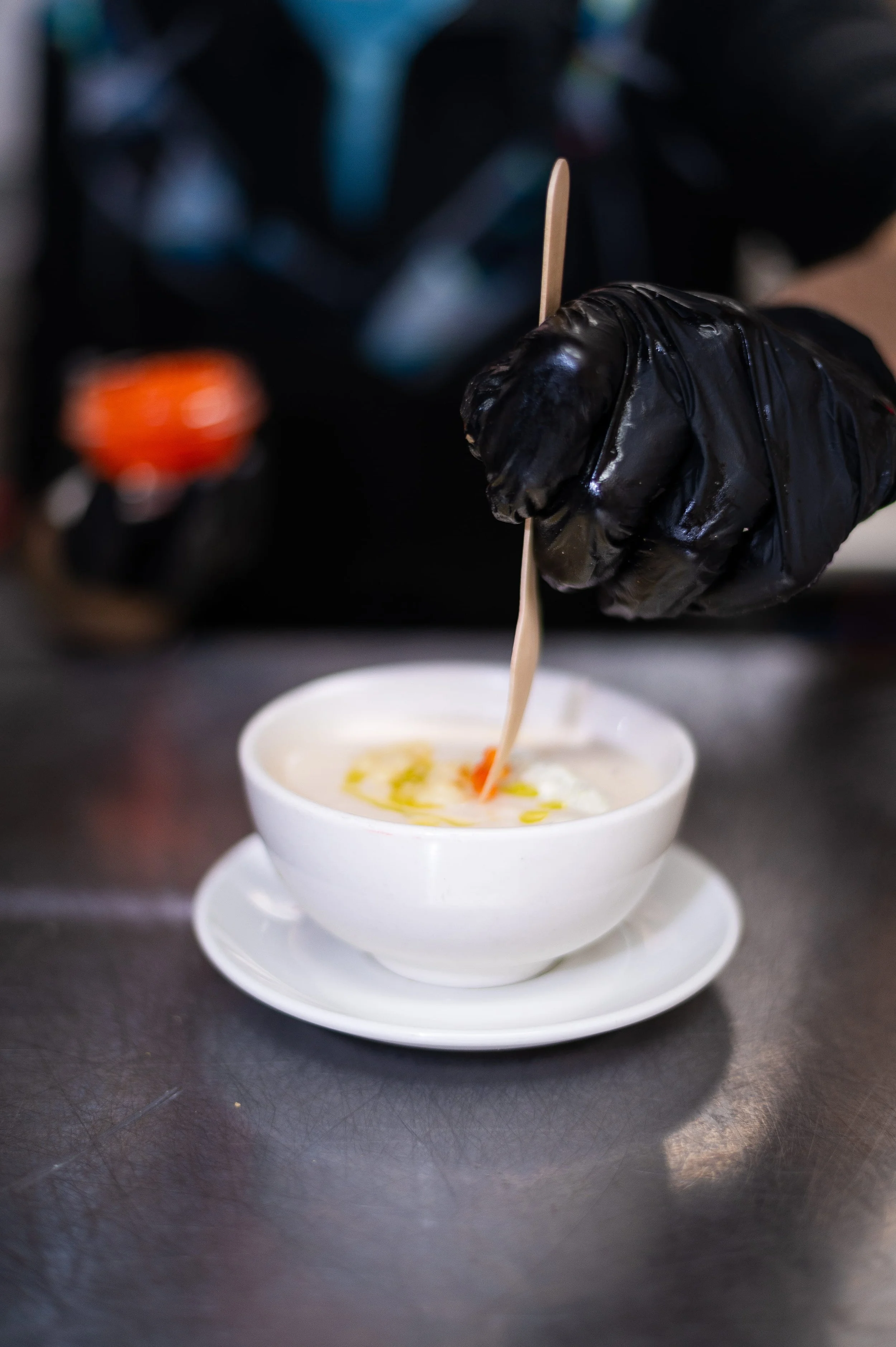 A gloved hand of the chef is holding a wooden stir stick over a bowl of soup with colorful toppings.