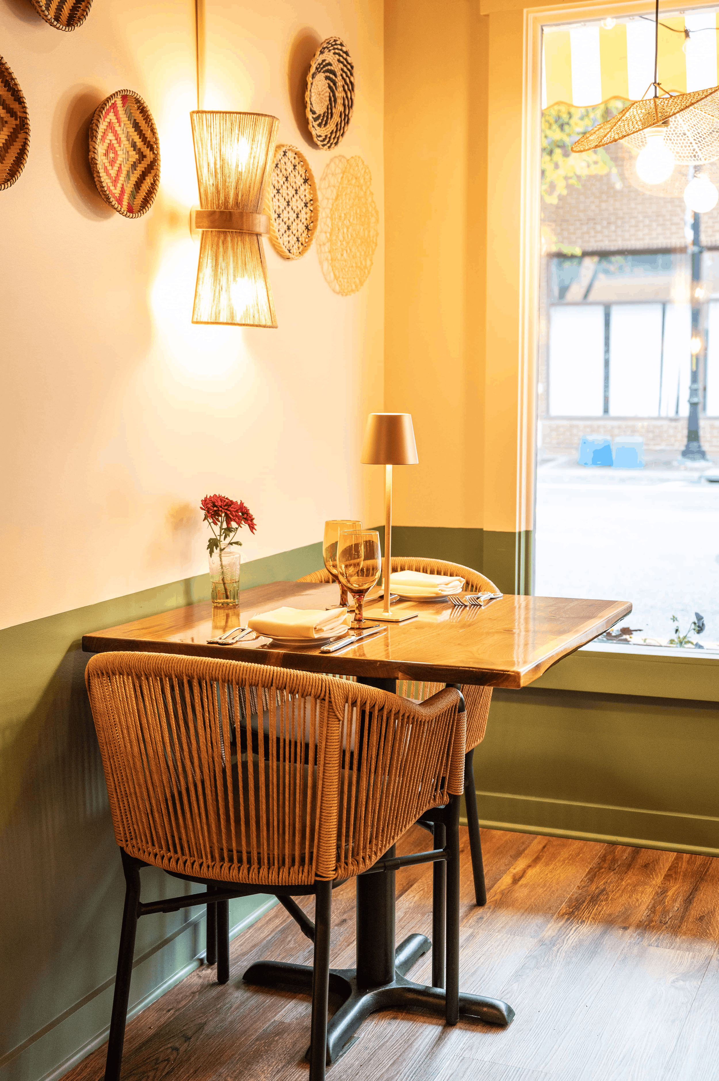 A cozy dining area with a wooden table set for two, including wine glasses, plates, and napkins. A small vase with pink flowers decorates the table. The wall behind features woven wall art and a wall-mounted light. A large window shows an outdoor scene with a brick building and trees, and the natural light streams into the room. Wicker chairs surround the table, and the floor is made of dark wood.