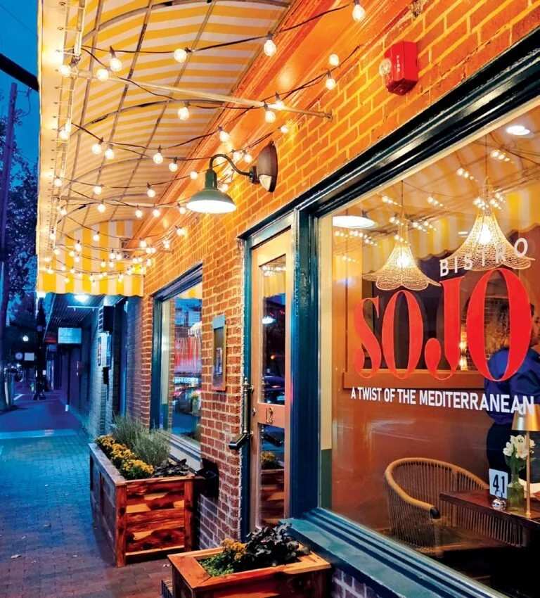 Exterior view of a restaurant with string lights hanging from a yellow and white striped awning, brick walls, large windows, and wooden planters with flowers on the sidewalk. The restaurant's window displays the name 'SOLO' with the tagline 'A Twist of the Mediterranean.'