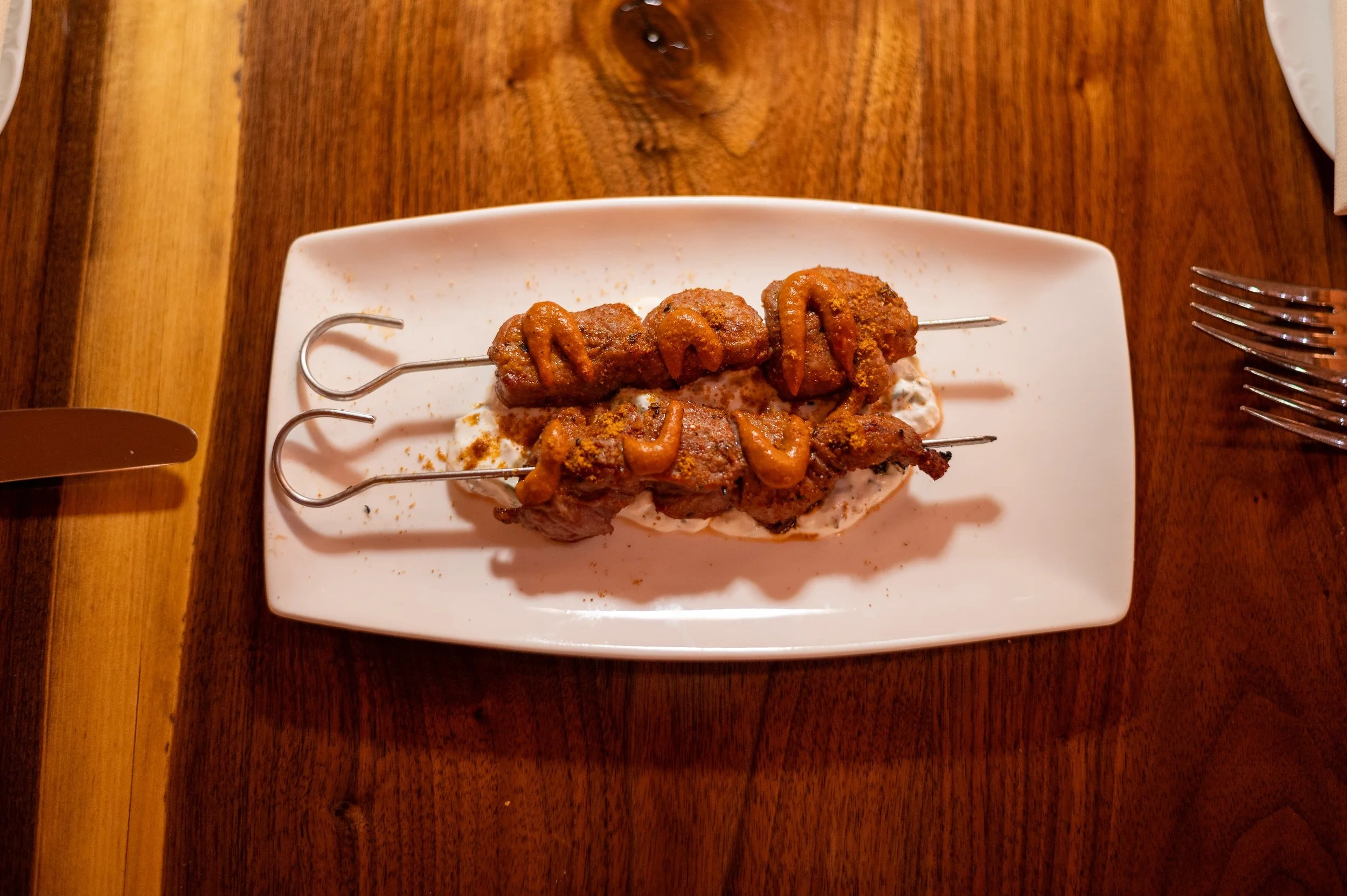 Three skewers of grilled meat or kebabs on a white rectangular plate, garnished with spices and sauce, served on a wooden table.