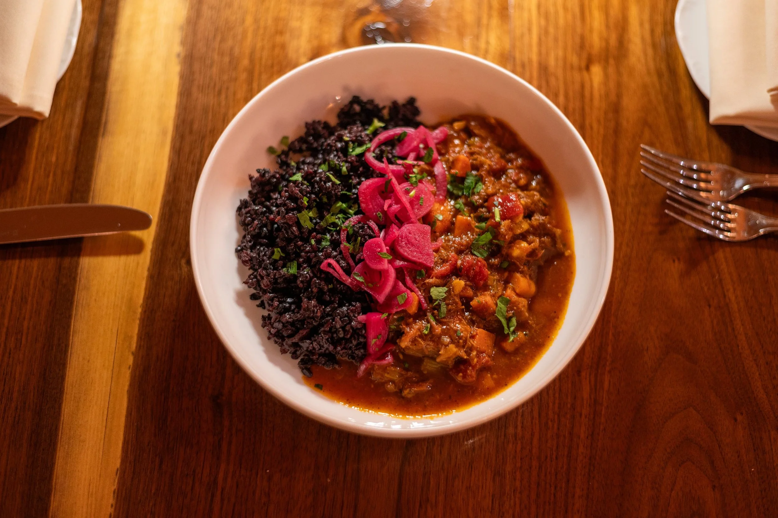 Mezze dish with rice and chili with pickled red onions, chopped herbs, served on a wooden table with cutlery and napkins.