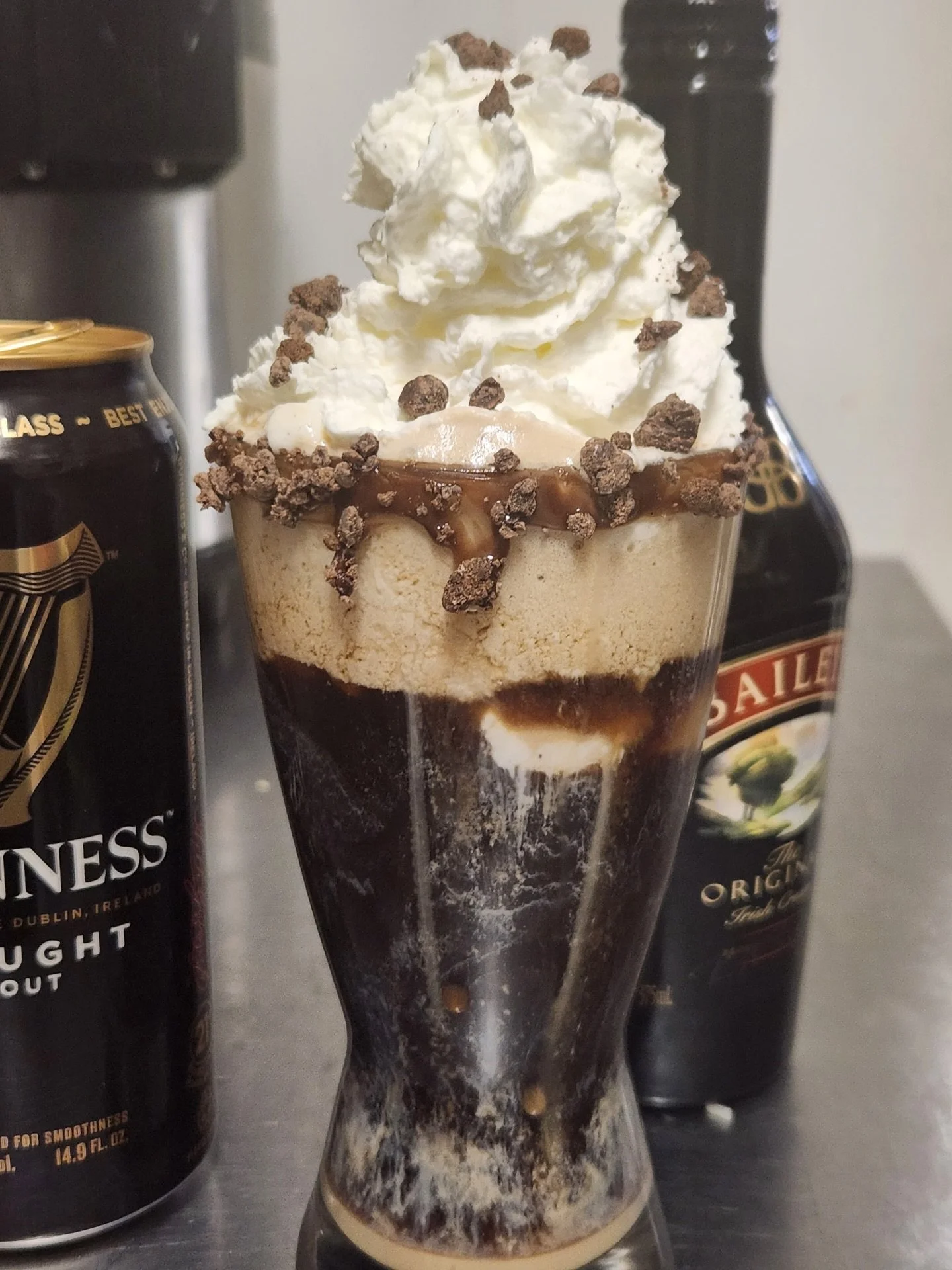A tall glass of layered coffee dessert topped with whipped cream, chocolate cookie crumbles, and caramel drizzle, surrounded by Guinness and Baileys Irish Cream bottles.