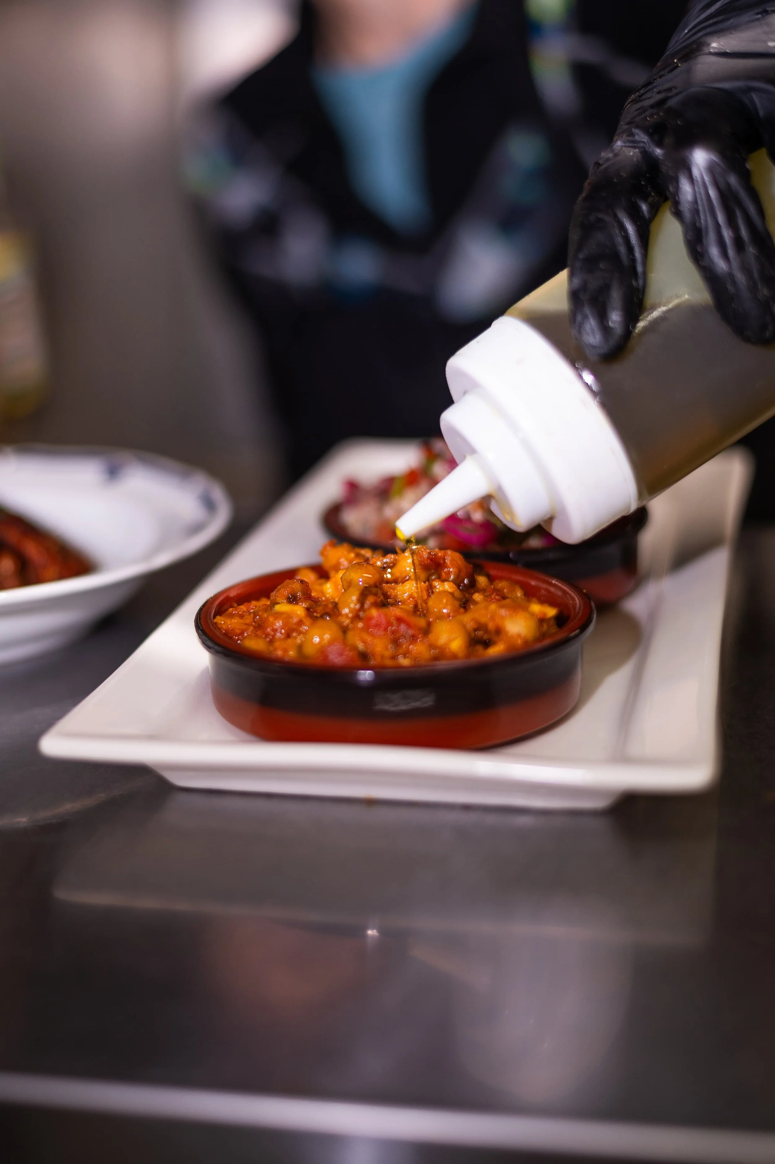 Chef wearing black gloves is squeezing signature sauce from a bottle into mezze bowls, with a blurred background of other food dishes.