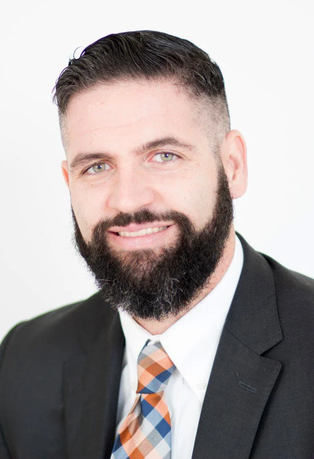 Headshot of a man, Of Counsel Patrick M. Coughlin,  with short dark hair, a full beard, light-colored eyes, wearing a dark suit jacket, white shirt, and a patterned tie, smiling against a plain white background.