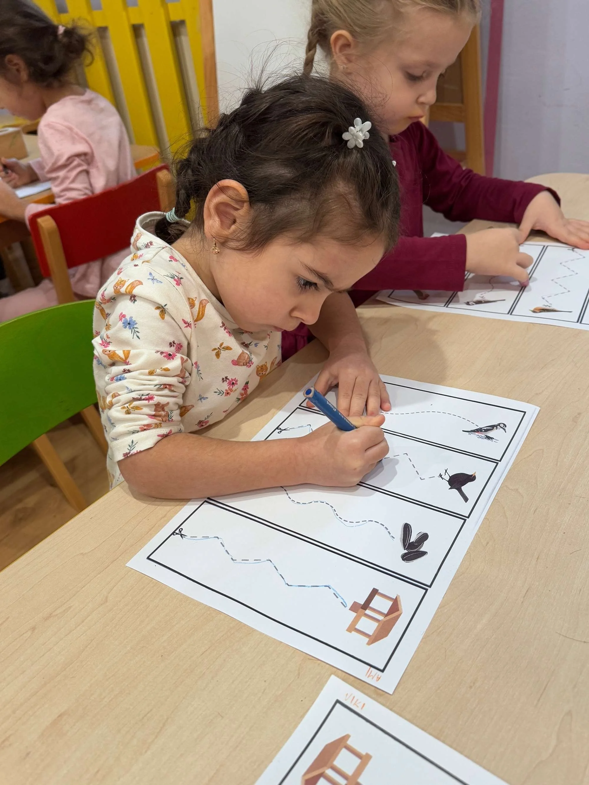 Young girl with dark hair in pigtails and a floral shirt, sitting at a table, coloring a worksheet with a blue marker. Another girl with blonde hair is sitting next to her, also working on a worksheet. There are children in the background, and the se