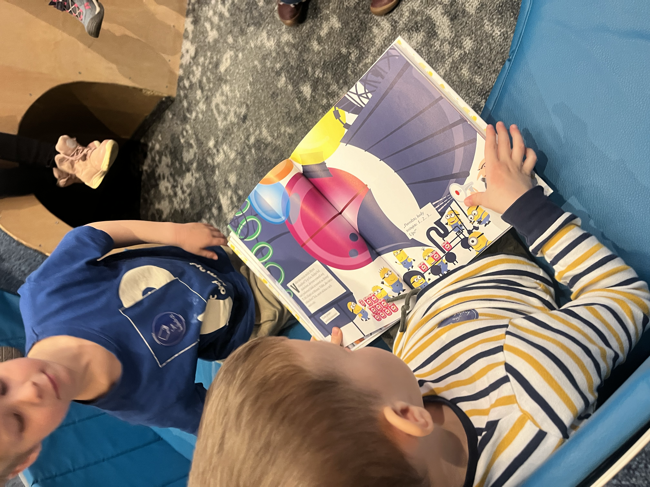 Two boys are sitting at a table, looking at a colorful cartoon magazine with cartoon characters and some numbers on the pages.