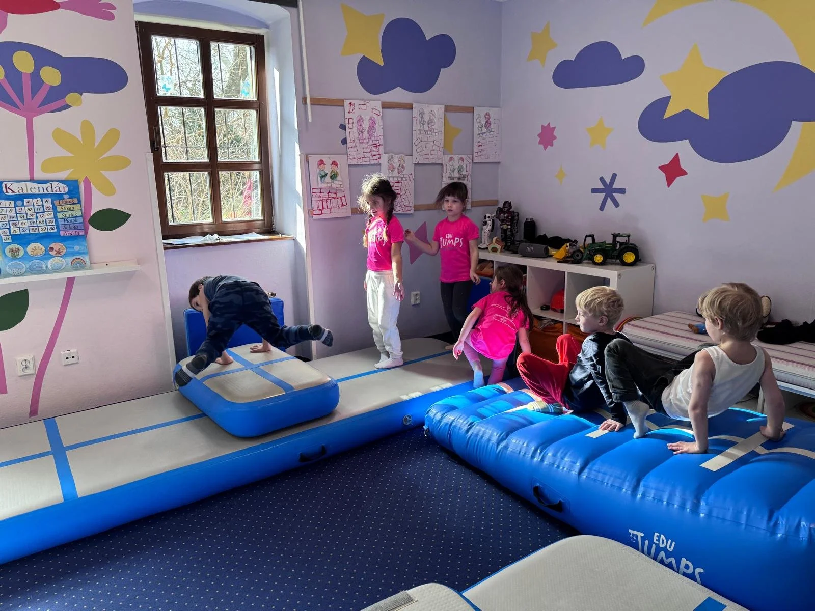Children playing and observing in a colorful playroom with cloud and star wall decorations. One child is climbing on an inflatable gym mat, while others are sitting or standing nearby.