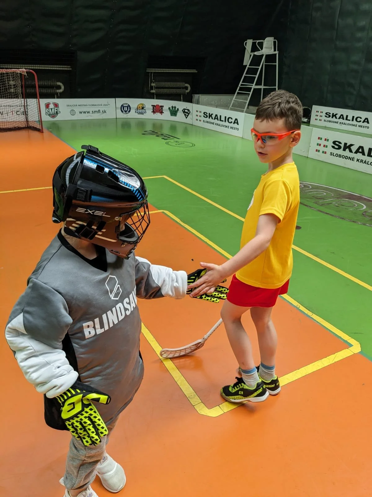 Two young boys on an indoor hockey rink, one dressed in goalie gear and helmet, the other in a yellow t-shirt and red shorts, shaking hands.