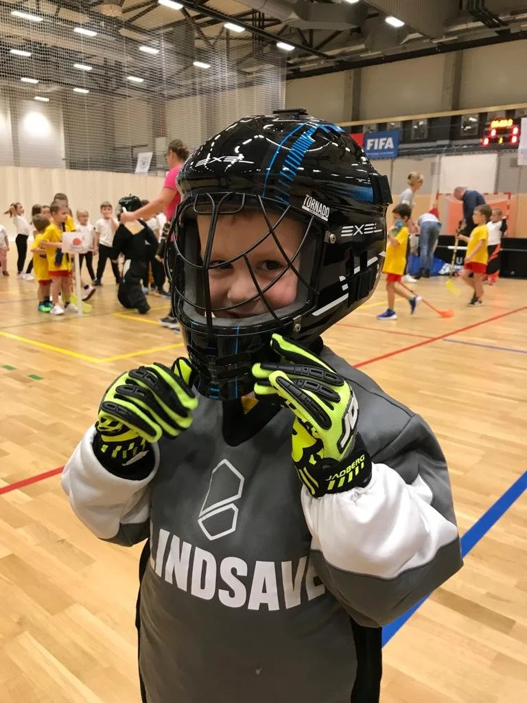 A young child dressed in hockey gear, including a helmet, gloves, and a jersey with 'HUNDSAVE' on it, standing on an indoor sports court with other children and adults in the background.