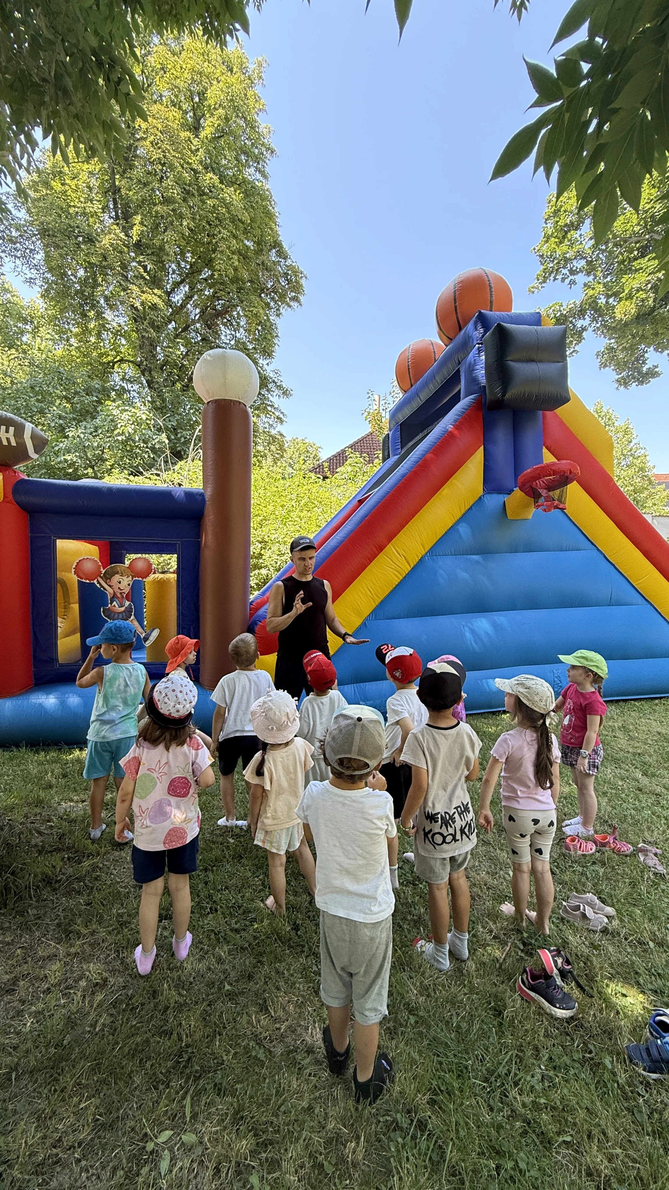 A group of children gathered around an outdoor inflatable bounce house with basketball hoops, under a clear sky and surrounded by green trees.
