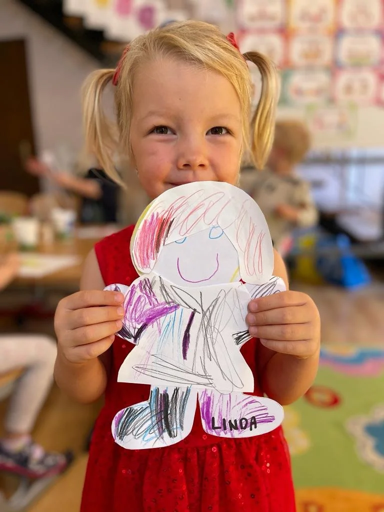 A young girl with blonde hair in pigtails, wearing a red dress, holding a colorful drawing of a person she made, with the name 'Linda' written on it, in a classroom setting.