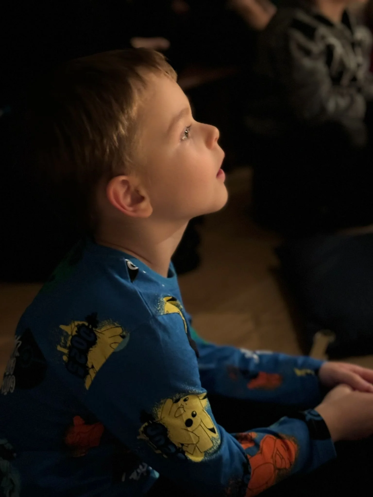 A young boy with short blond hair wearing colorful pajamas featuring characters from Pokémon, sitting in a dark room, attentively watching something with a focused expression.