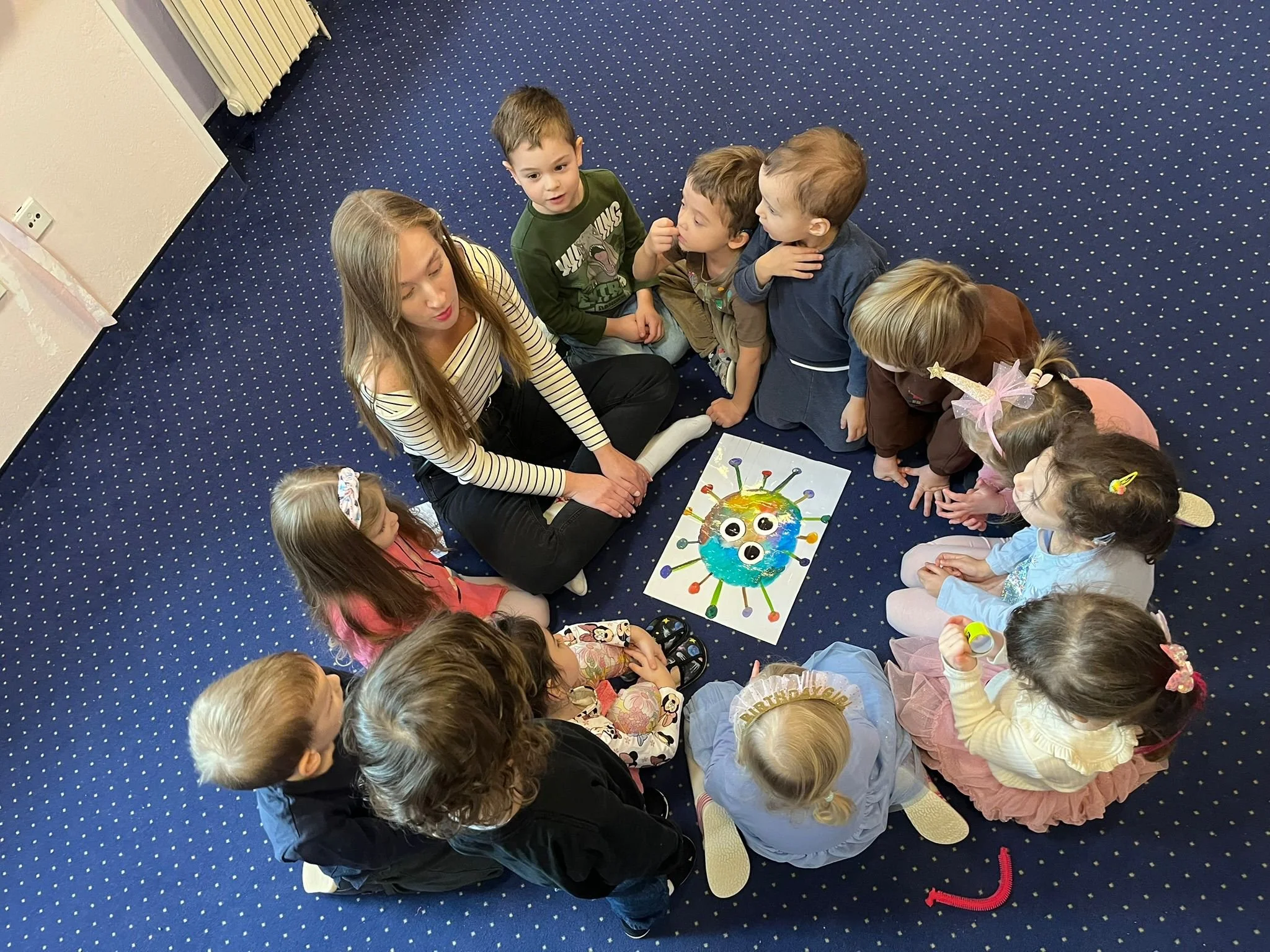 A group of children and a woman sitting in a circle on a blue carpet with white dots, gathered around a colorful illustrated drawing of a virus with googly eyes and raised antennae.