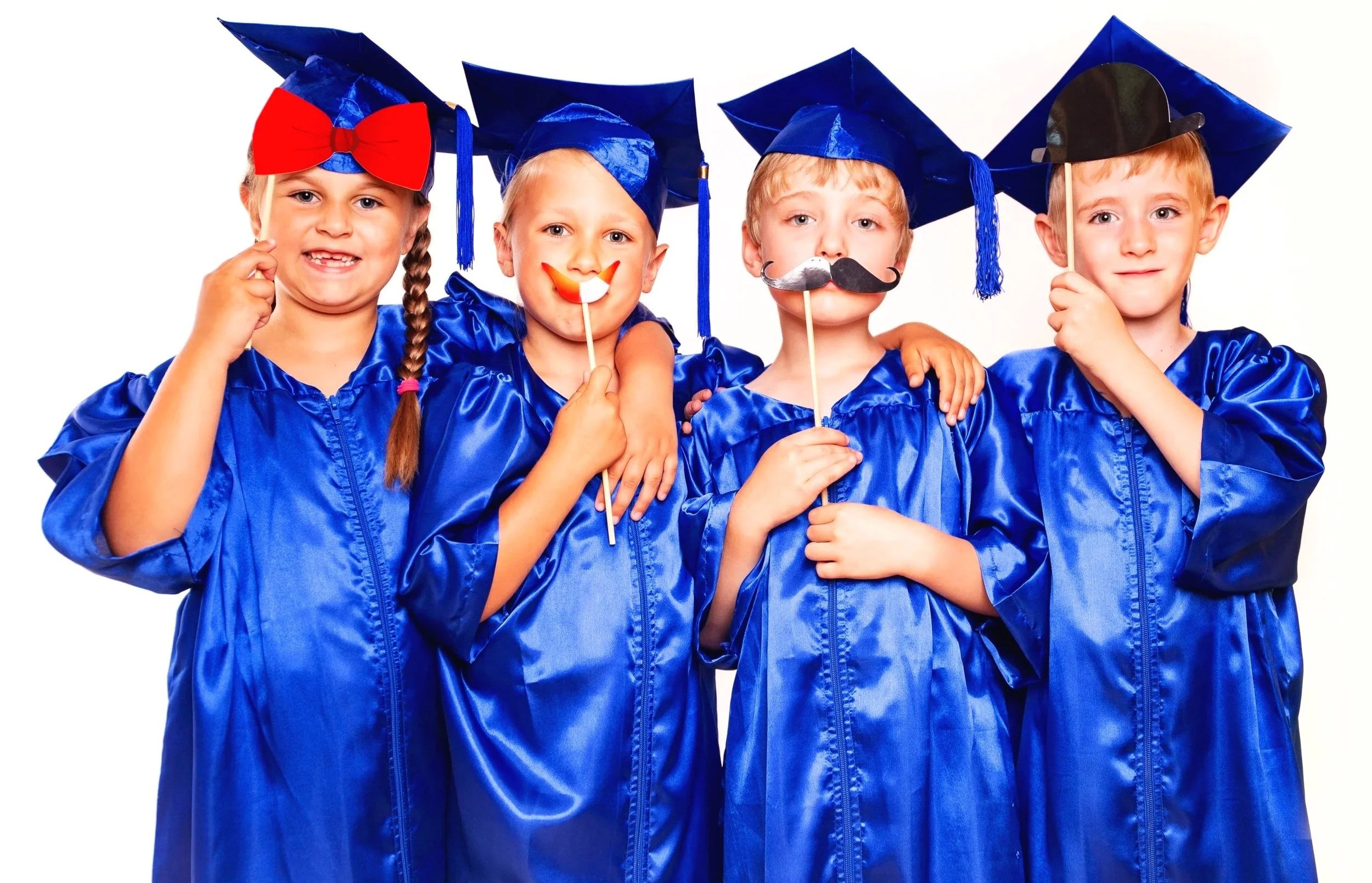 Four children in blue graduation gowns and caps, holding playful props like a big red bow, a smiley face, a mustache, and a baseball cap, smiling against a white background.