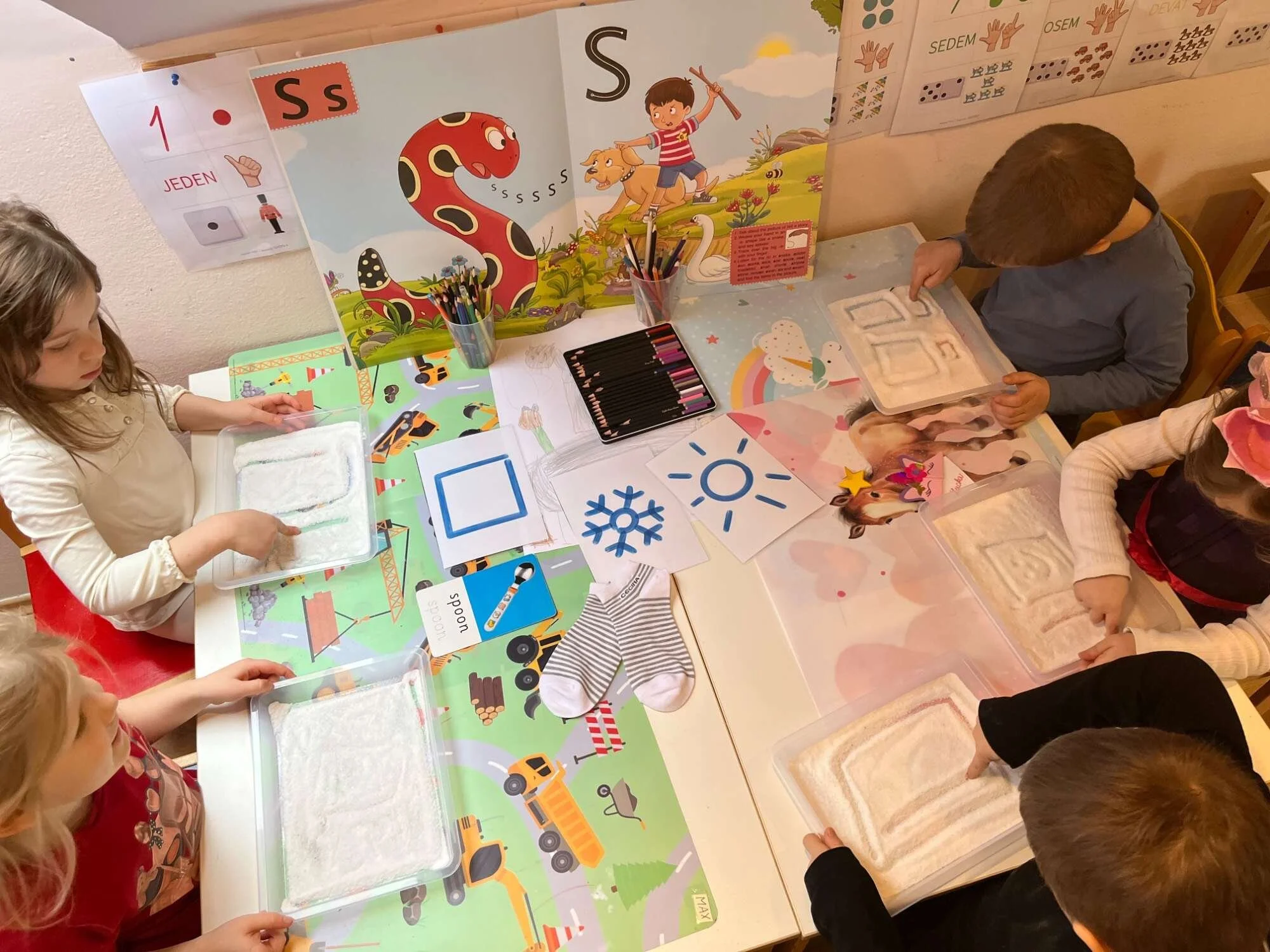 Five children sitting around a Christmas-themed table, engaging in a sensory activity with trays of white sand or salt, with colorful posters, markers, and children's drawings on the wall and table.