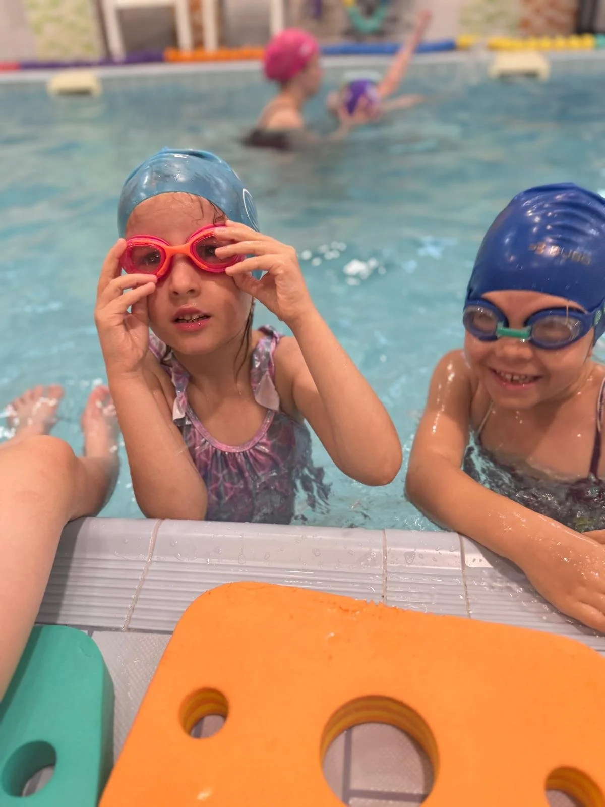 Two children in swimming caps and goggles at a pool, with another person and kids swimming in the background.
