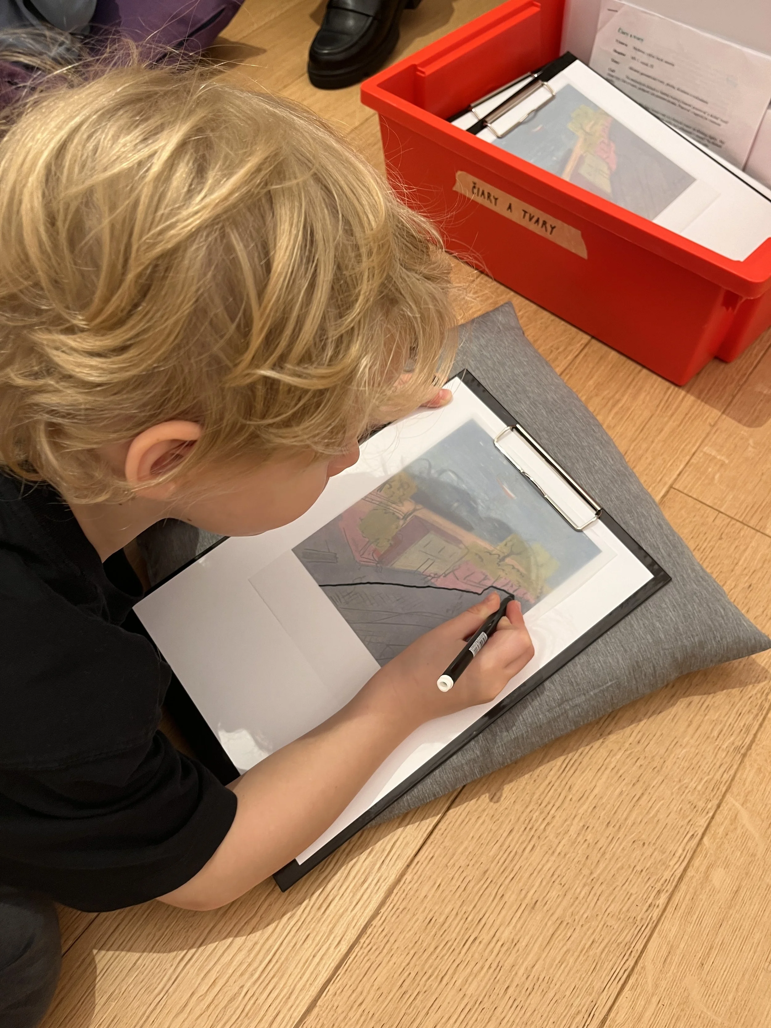 A young child with blond hair drawing on a clipboard with a black marker, sitting on a wooden floor. There is a red box labeled 'CHIART A TVARY' containing papers and pens nearby.