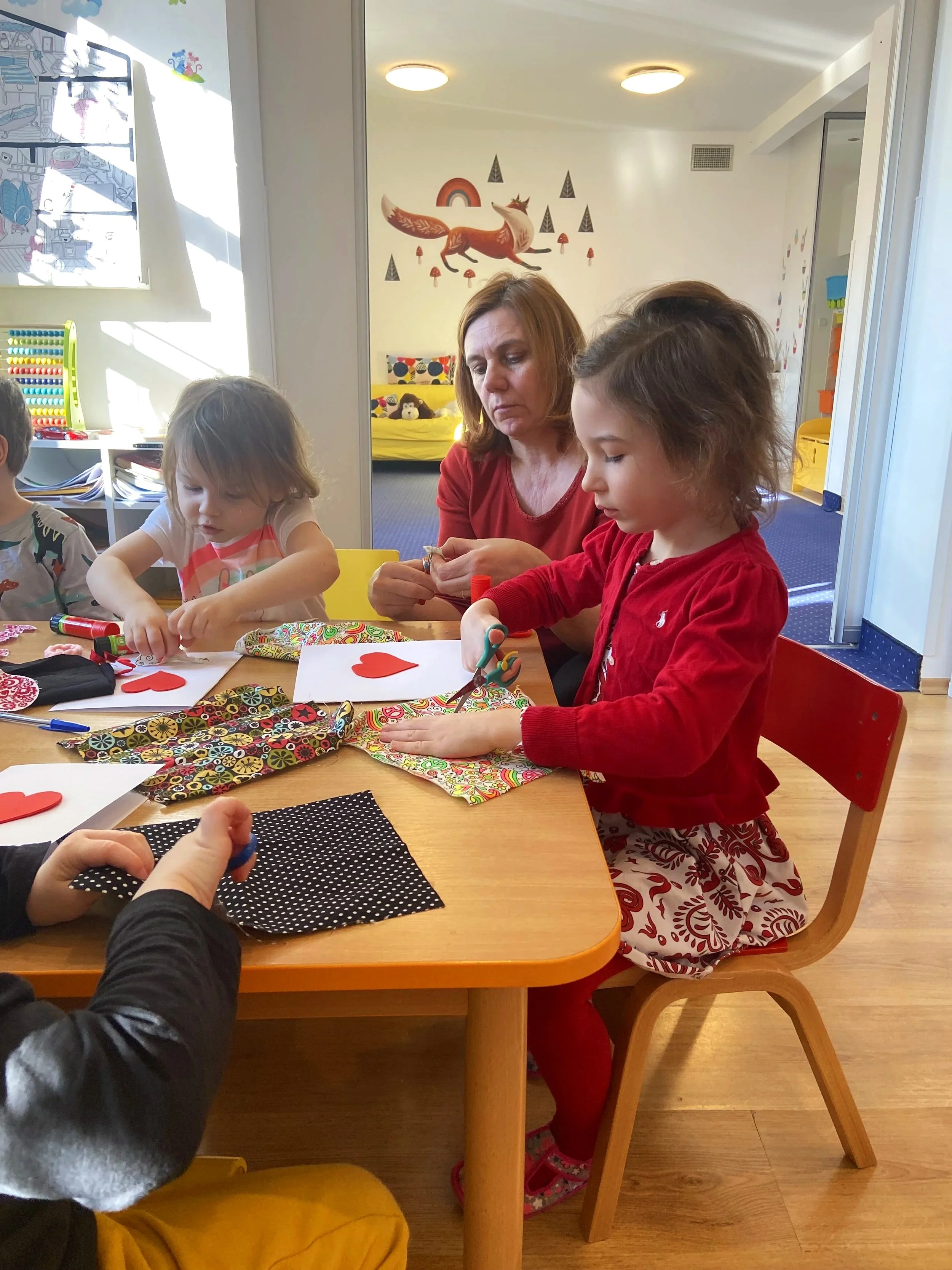 Children and adult sitting at a table engaging in arts and crafts, with decorating materials, paper hearts, scissors, and fabric on the table, in a brightly lit room with cheerful decorations on the walls.