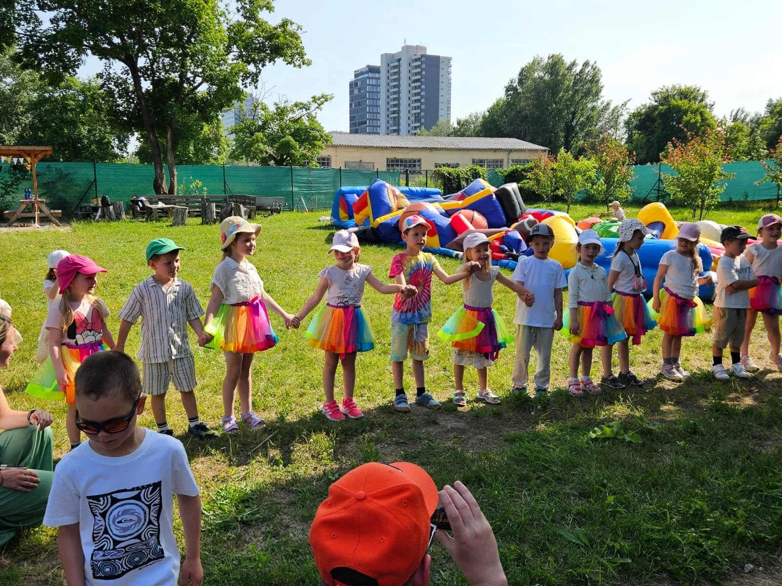 Children dressed in colorful skirts and white shirts participate in a line dance outdoors, with inflatable structures and trees in the background on a sunny day.
