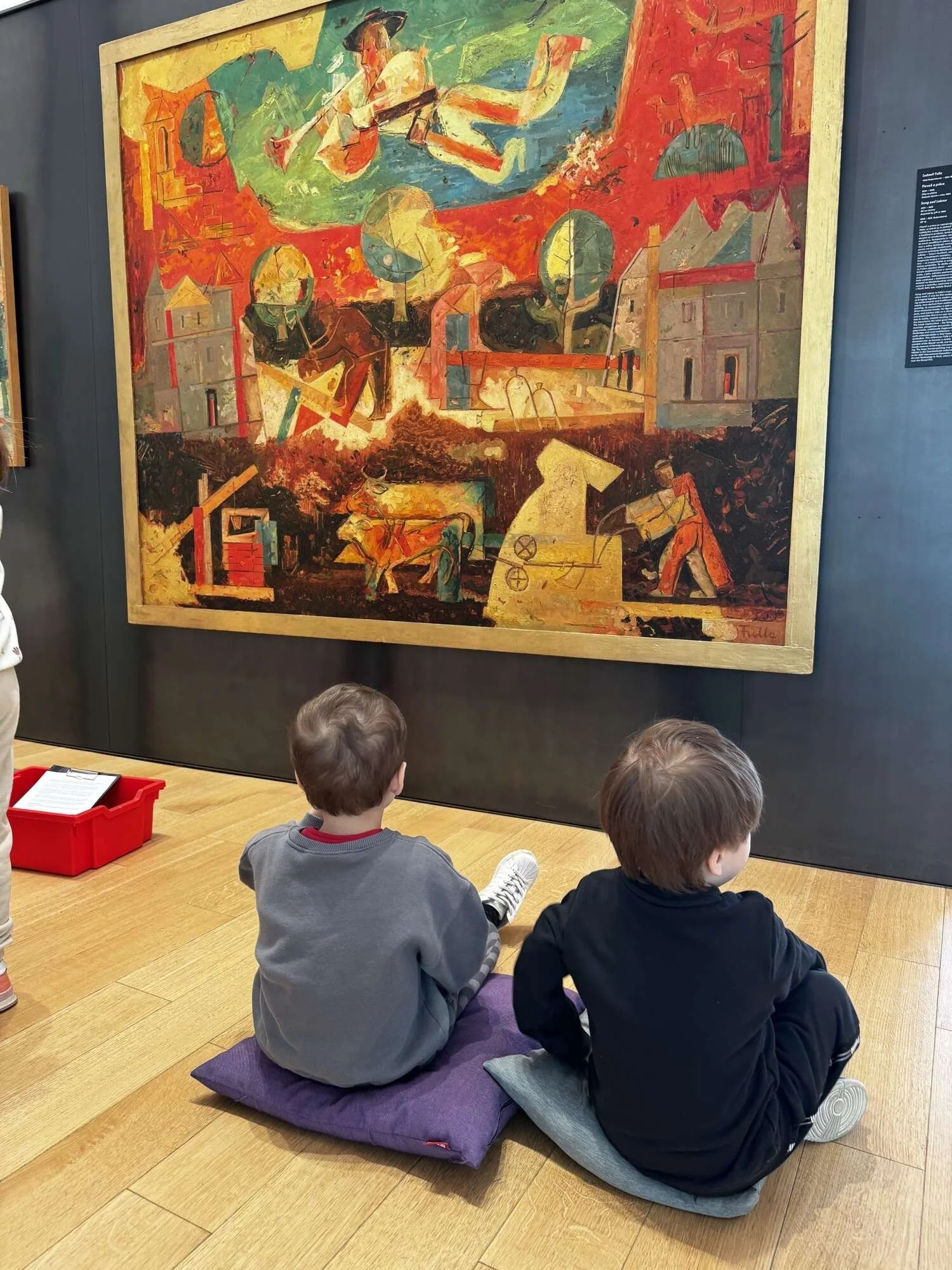 Two young boys sitting on cushions on the floor in front of a colorful, abstract painting in a museum.