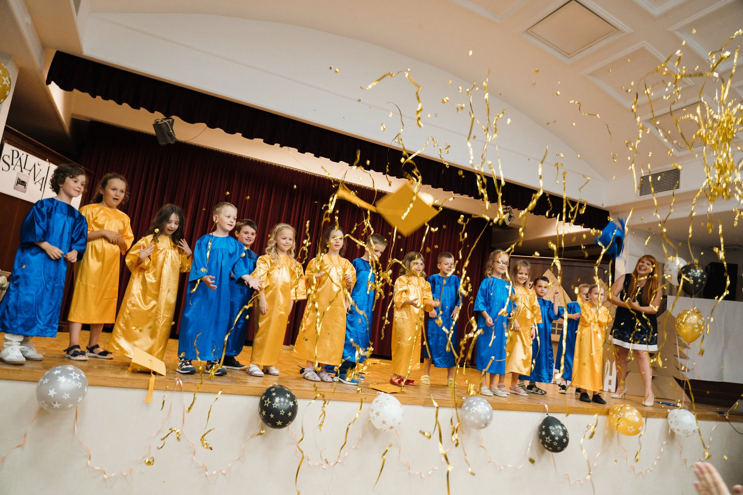 Children in graduation gowns on stage celebrating with balloons and confetti.
