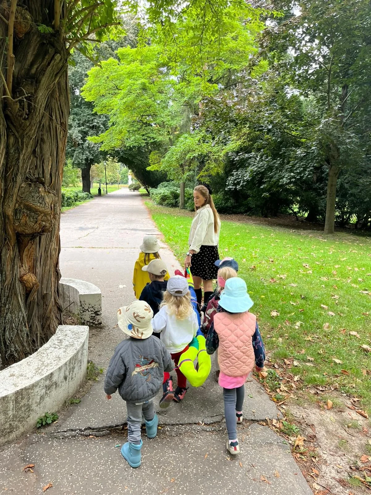 A woman leading a group of young children on a walk in a park, surrounded by green trees and grass.