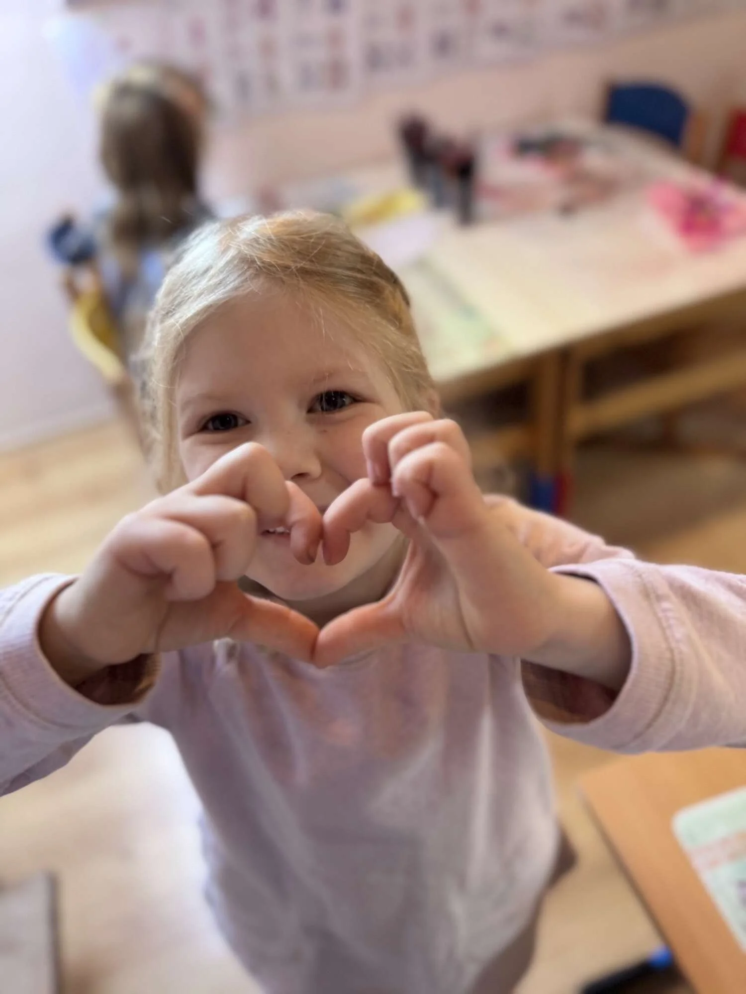 A young girl making a heart shape with her hands, smiling, in a classroom.