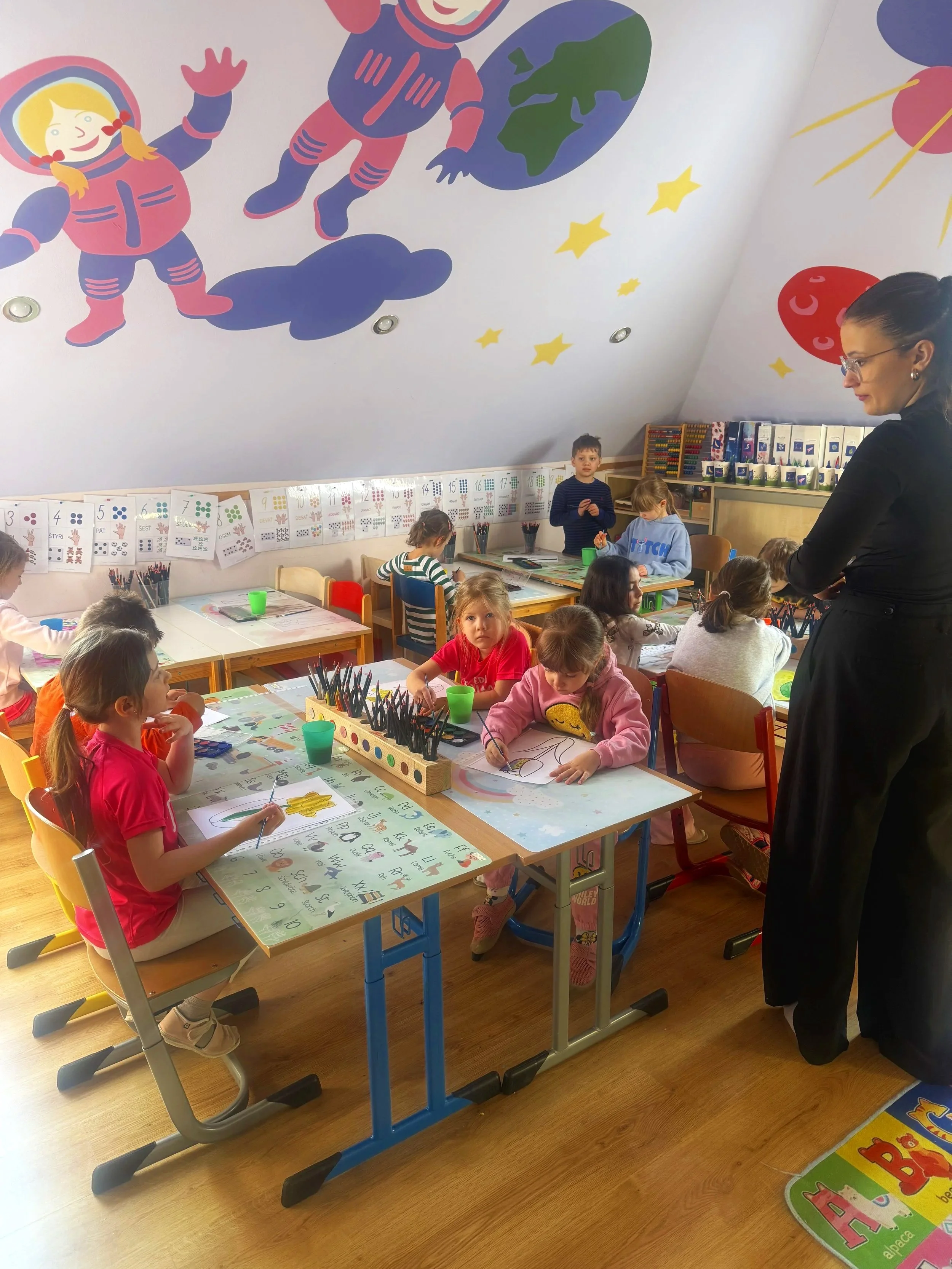 A classroom filled with young children working on drawings at desks. An adult woman stands observing. The ceiling has colorful space-themed murals with astronauts, planets, and stars.