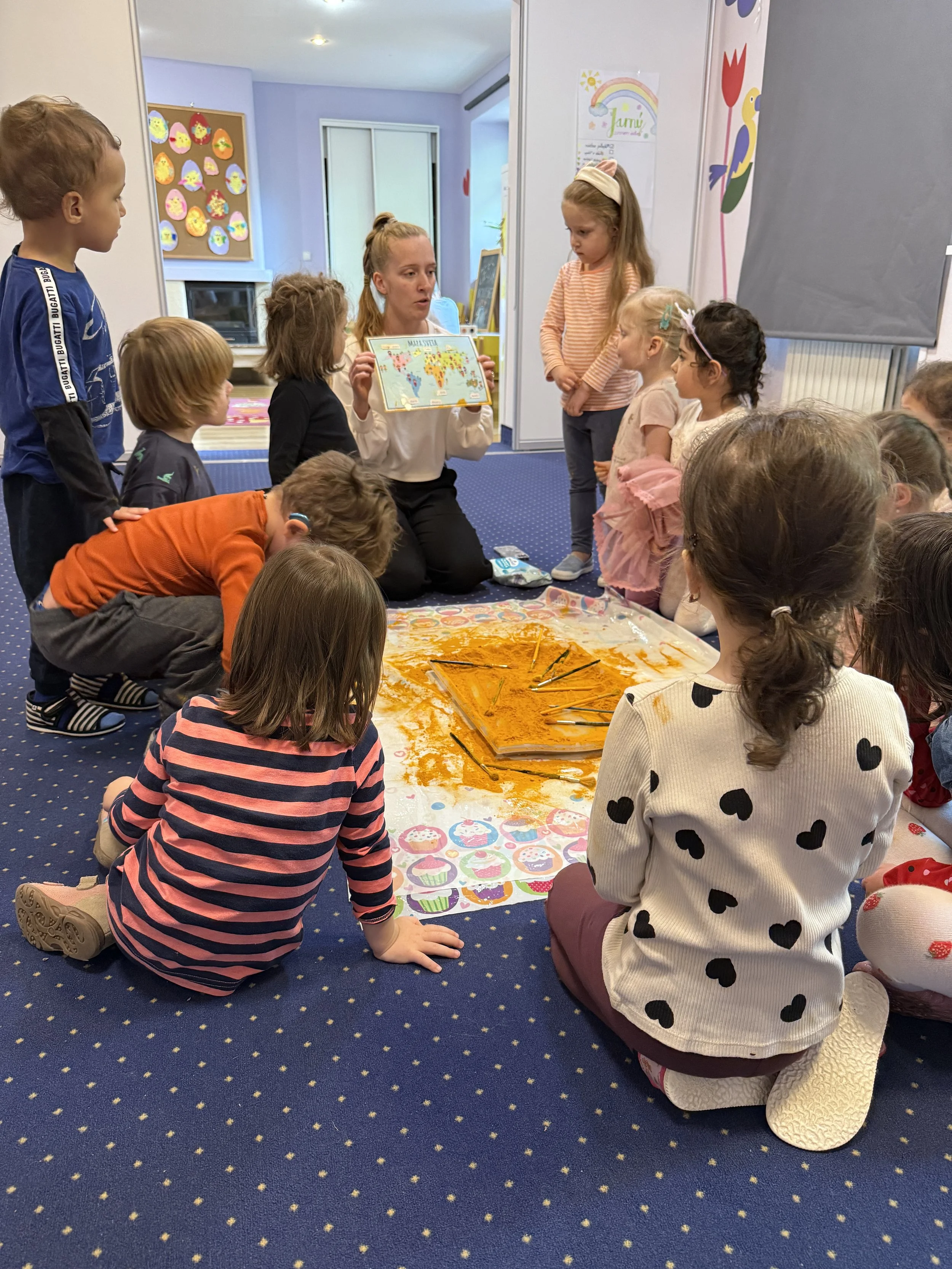 A teacher is kneeling on the carpet, holding a world map, explaining to a group of children gathered around her. The children are sitting or kneeling on a colorful, cupcake-patterned mat, listening attentively. The room has colorful decorations and c