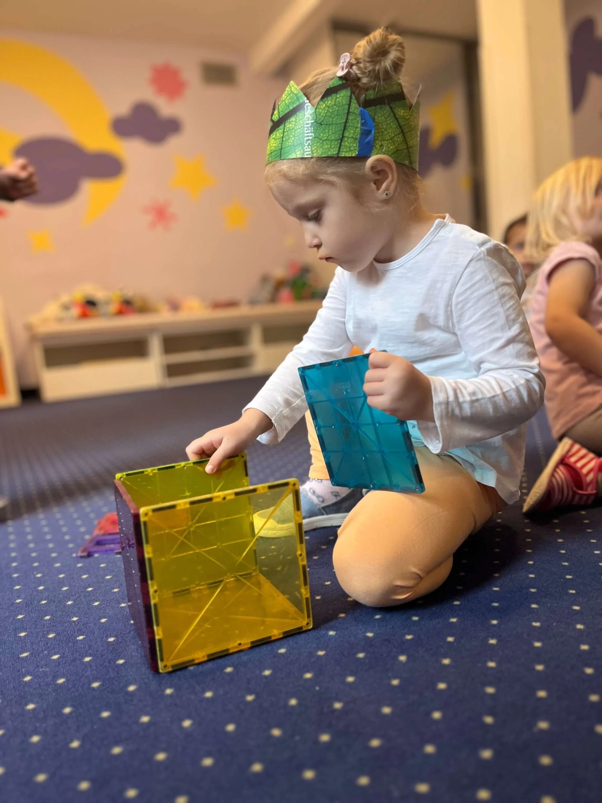 A young girl wearing a paper crown with green and blue designs, sitting on a navy blue carpet with yellow dots, is playing with colorful magnetic building blocks. The background shows a wall with cloud, star, and moon decorations.