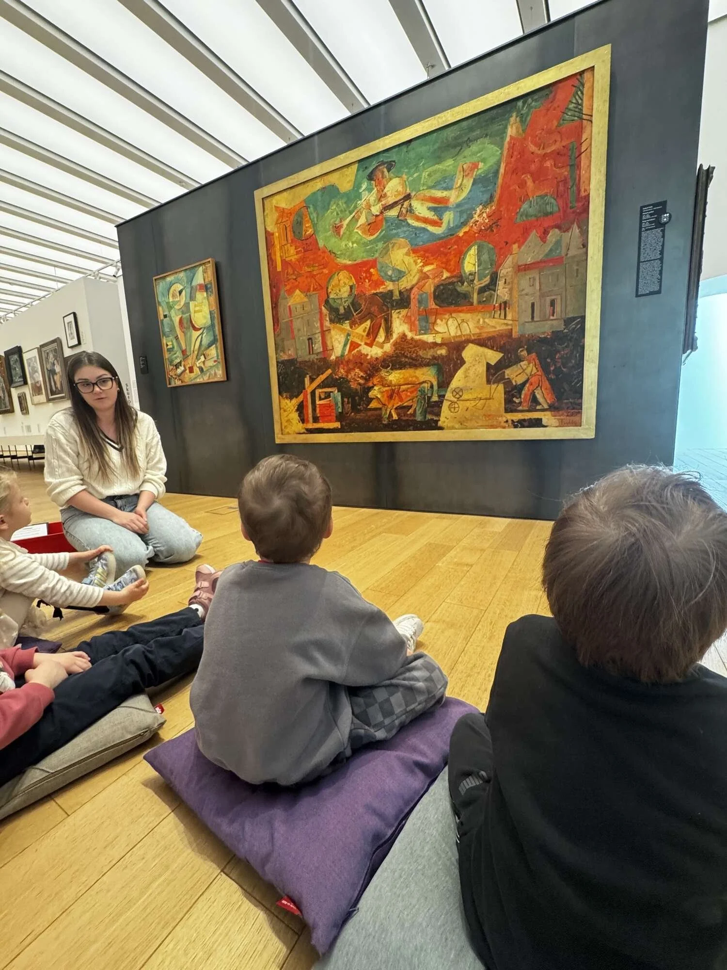 Children sitting on the floor and listening to a woman in a museum, with large colorful abstract paintings on the wall behind her.