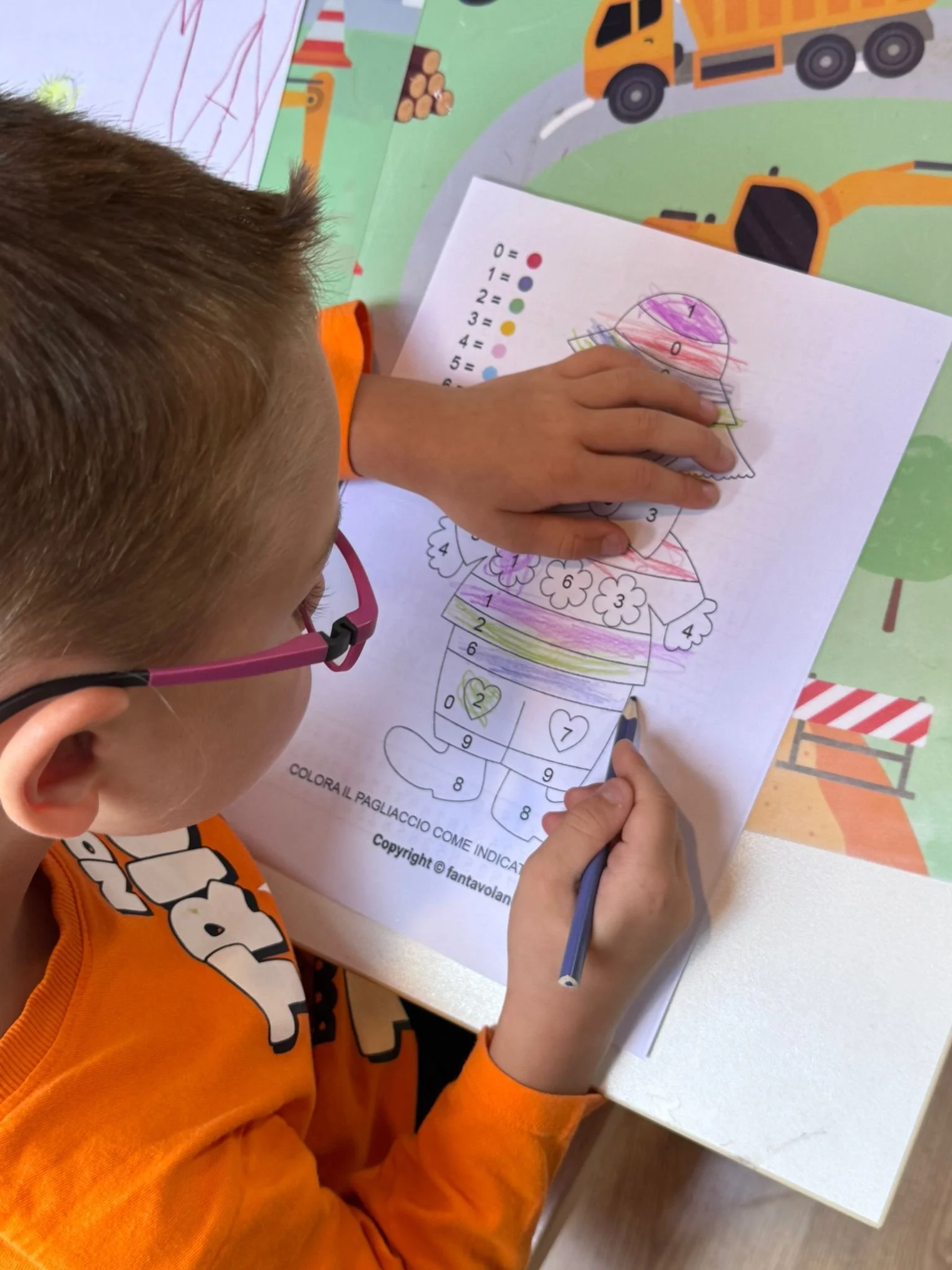 A young child wearing orange shirt and pink glasses coloring a numbered picture of a clown on a piece of paper with colored pencils. The child is working on a colorful craft activity at a table.