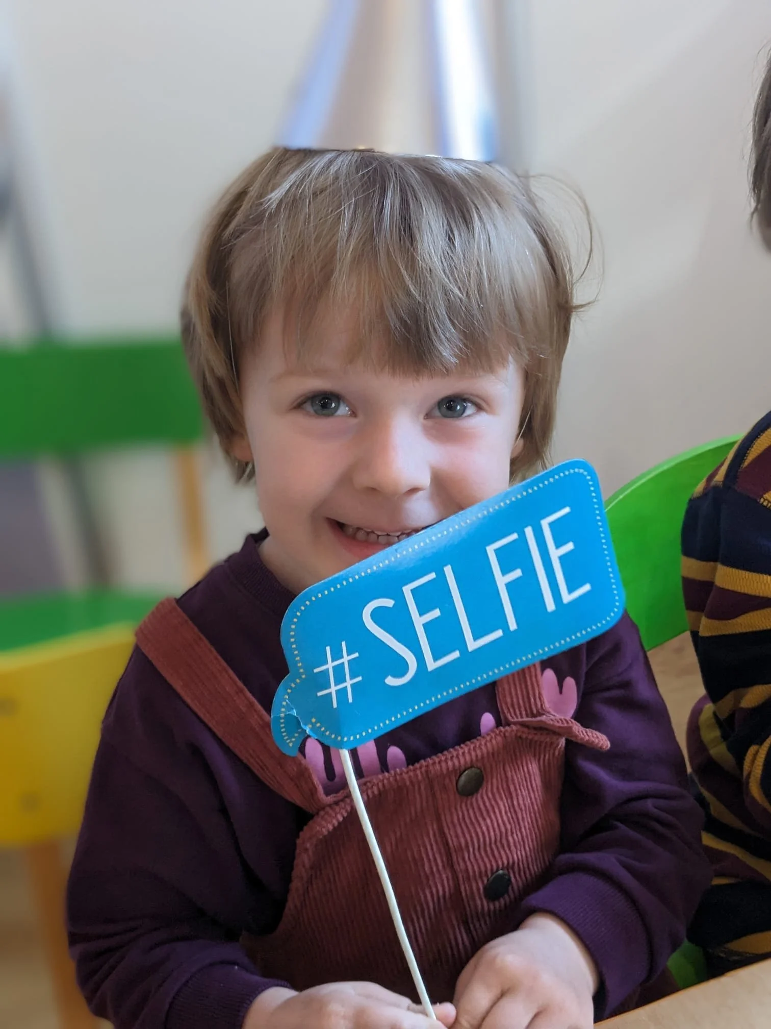 A young child with light brown hair and blue eyes smiles while holding a blue sign that says '#SELFIE'. The child is sitting at a table with colorful green and yellow chairs in the background.