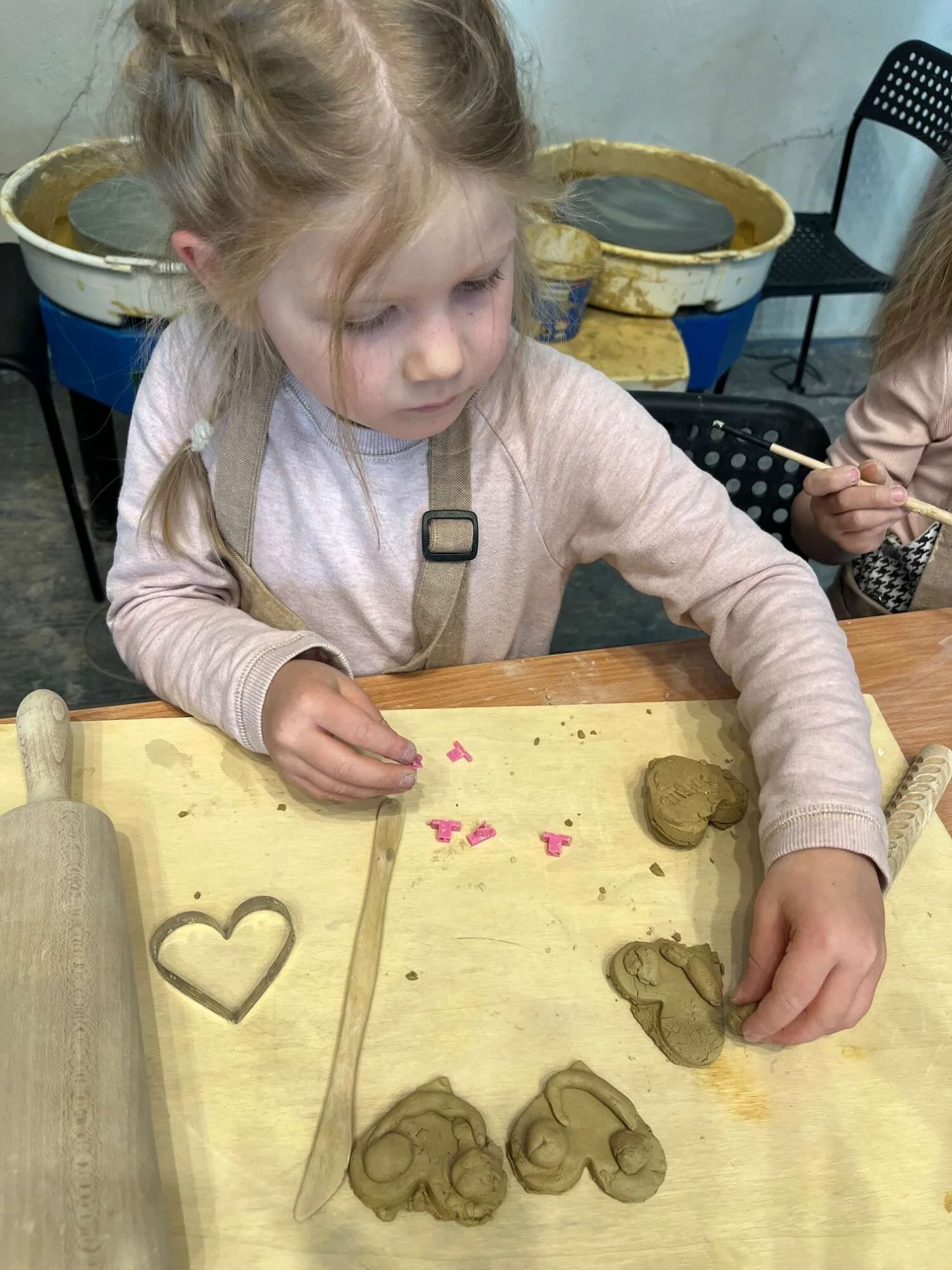 A young girl working with clay on a wooden table, surrounded by pottery tools and shapes.